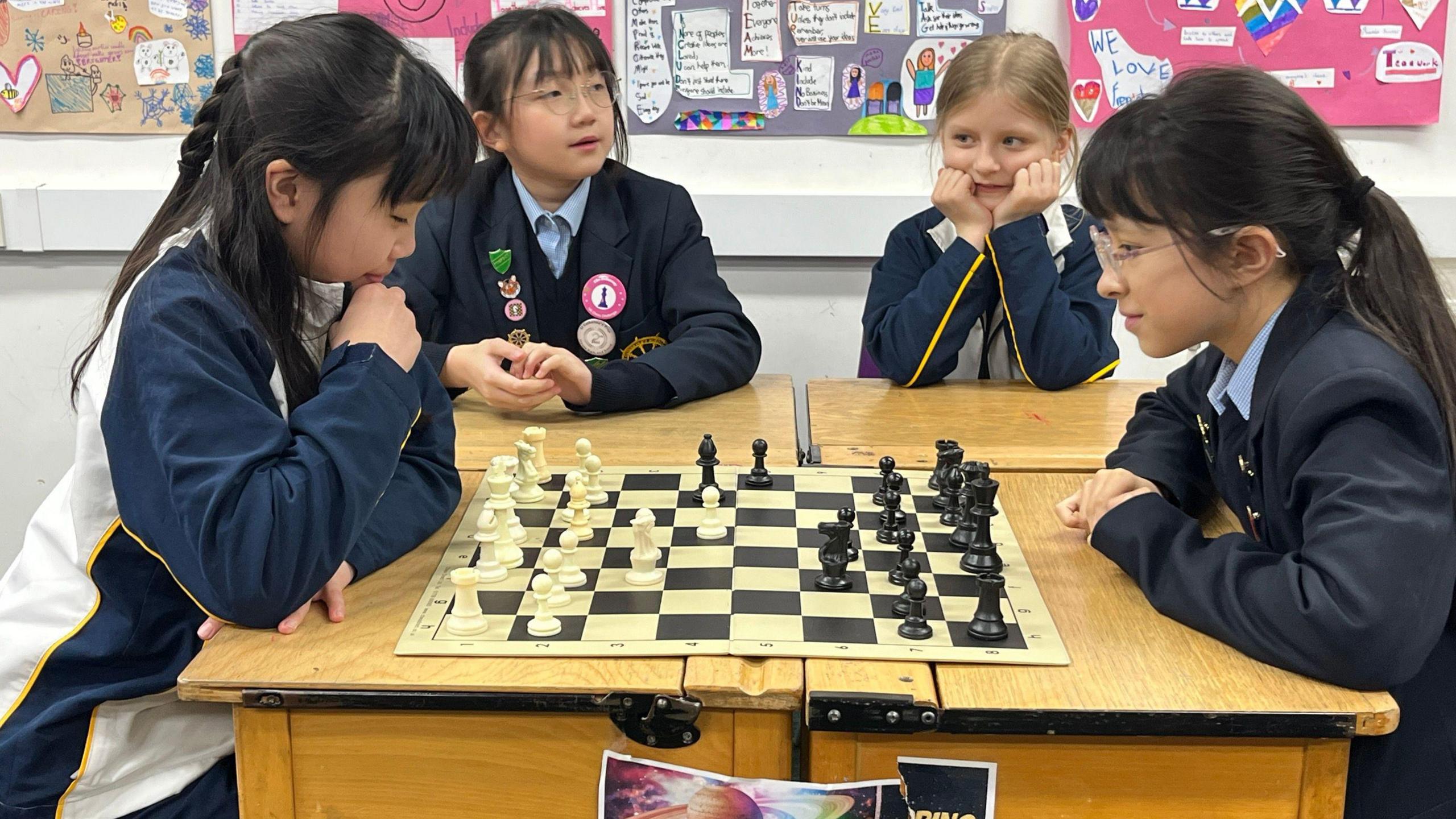 Two school girls are playing chess while another two school girls are sitting nearby.