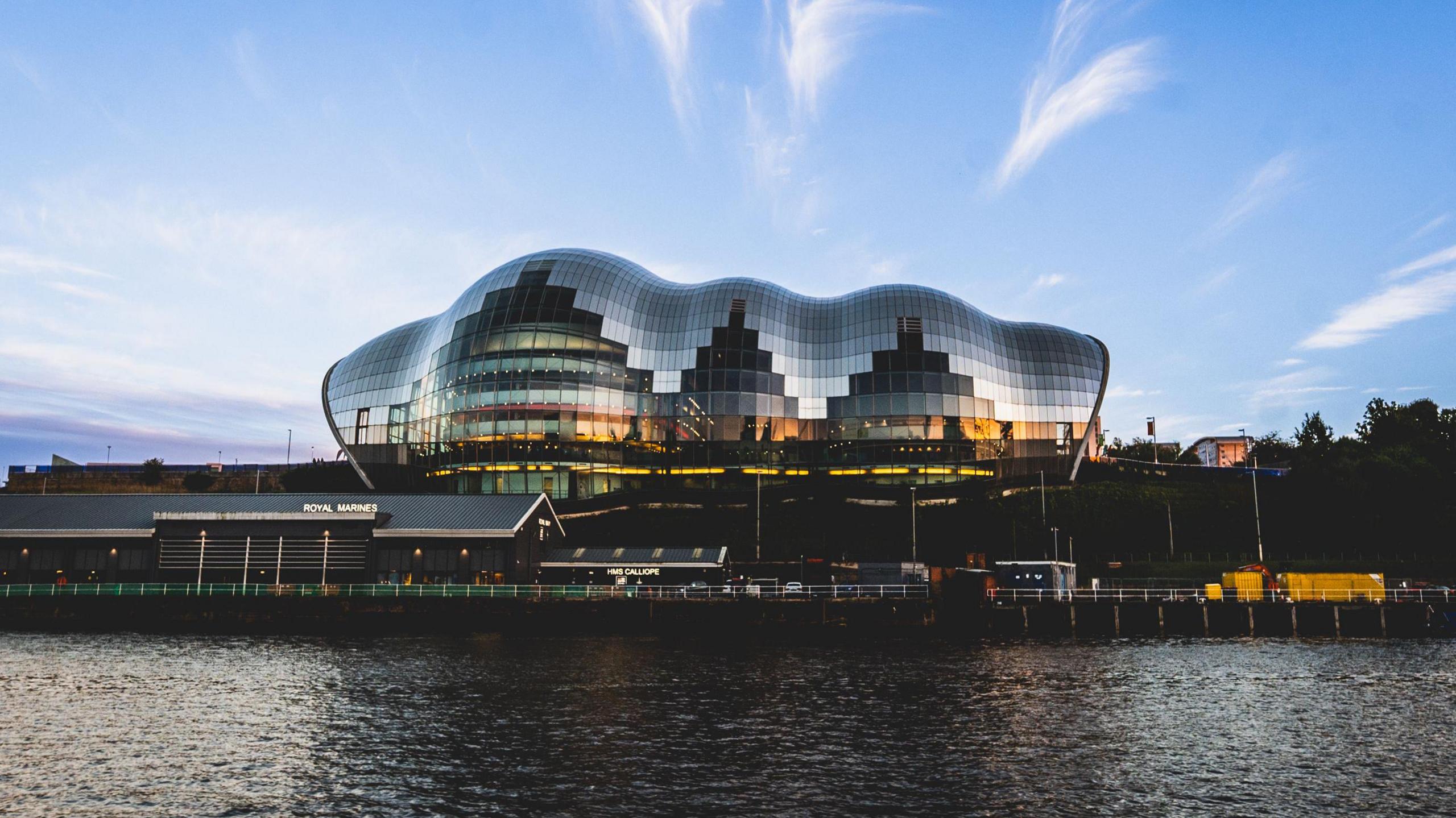 An image of the glasshouse international centre for music which has reflective panels on the outside. In the foreground are some smaller buildings and a river. In the background there's a blue sky with some light clouds. The photo has been taken at dusk so the light is fading.