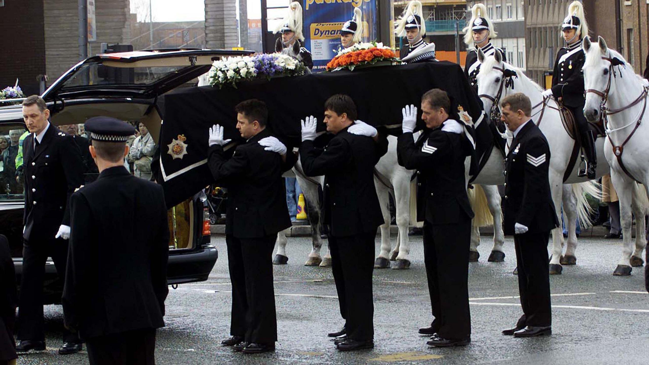The coffin of Detective Constable Stephen Oake is held by six pallbearers all in Greater Manchester Police uniform with white gloves. In the background mounted police officers have formed a guard of honour for the funeral cortege.