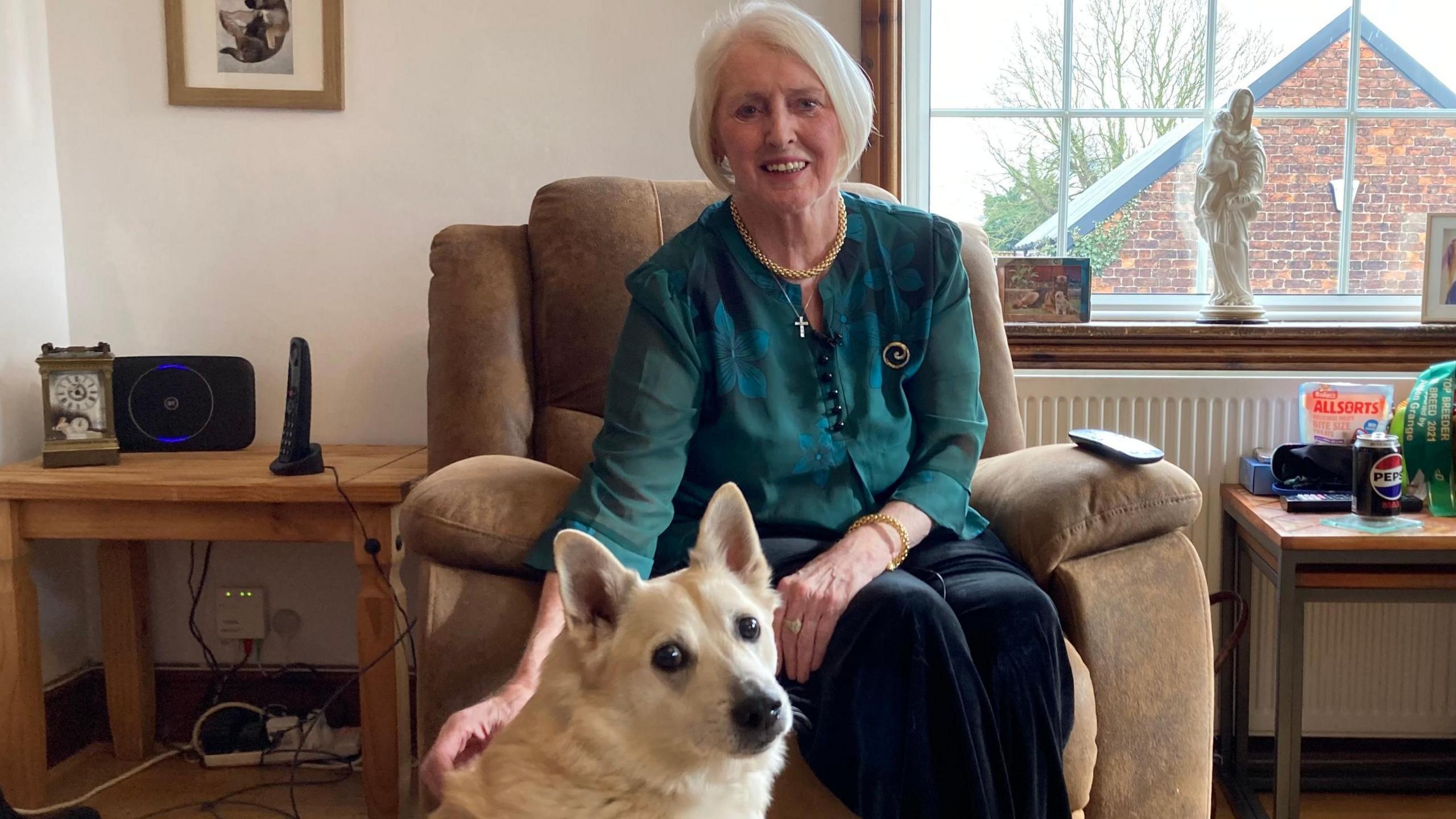 Diana Stirling has a grey/platinum-blonde short bob hairstyle and is wearing a dark green top. She is sitting in an armchair in her living room with her dog Sirius sitting at her feet looking towards the camera. Di is smiling at the camera.