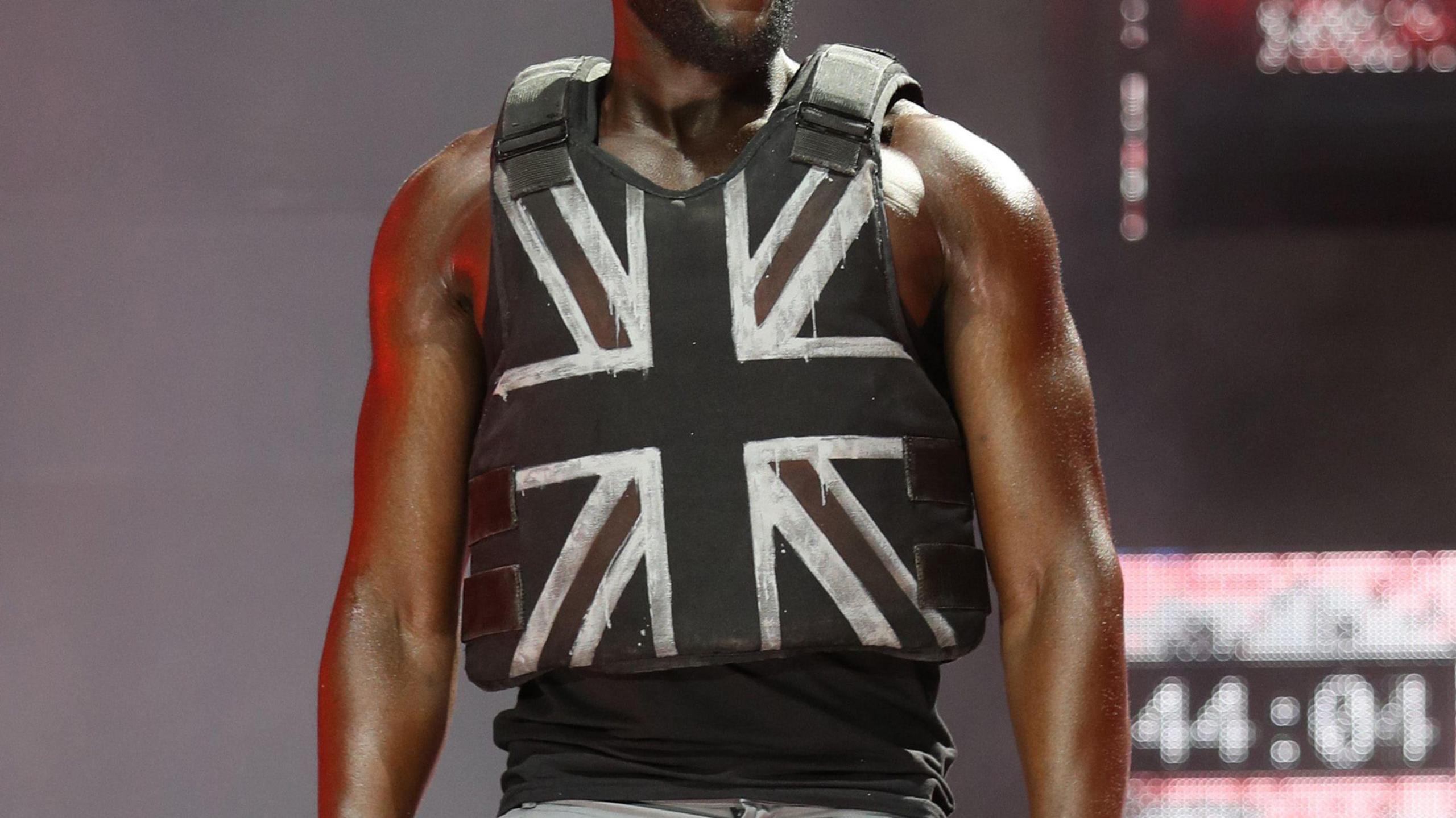 A close up of Stormzy on stage, wearing Banksy's bullet proof vest with a Union Jack flag spray painted onto it. 