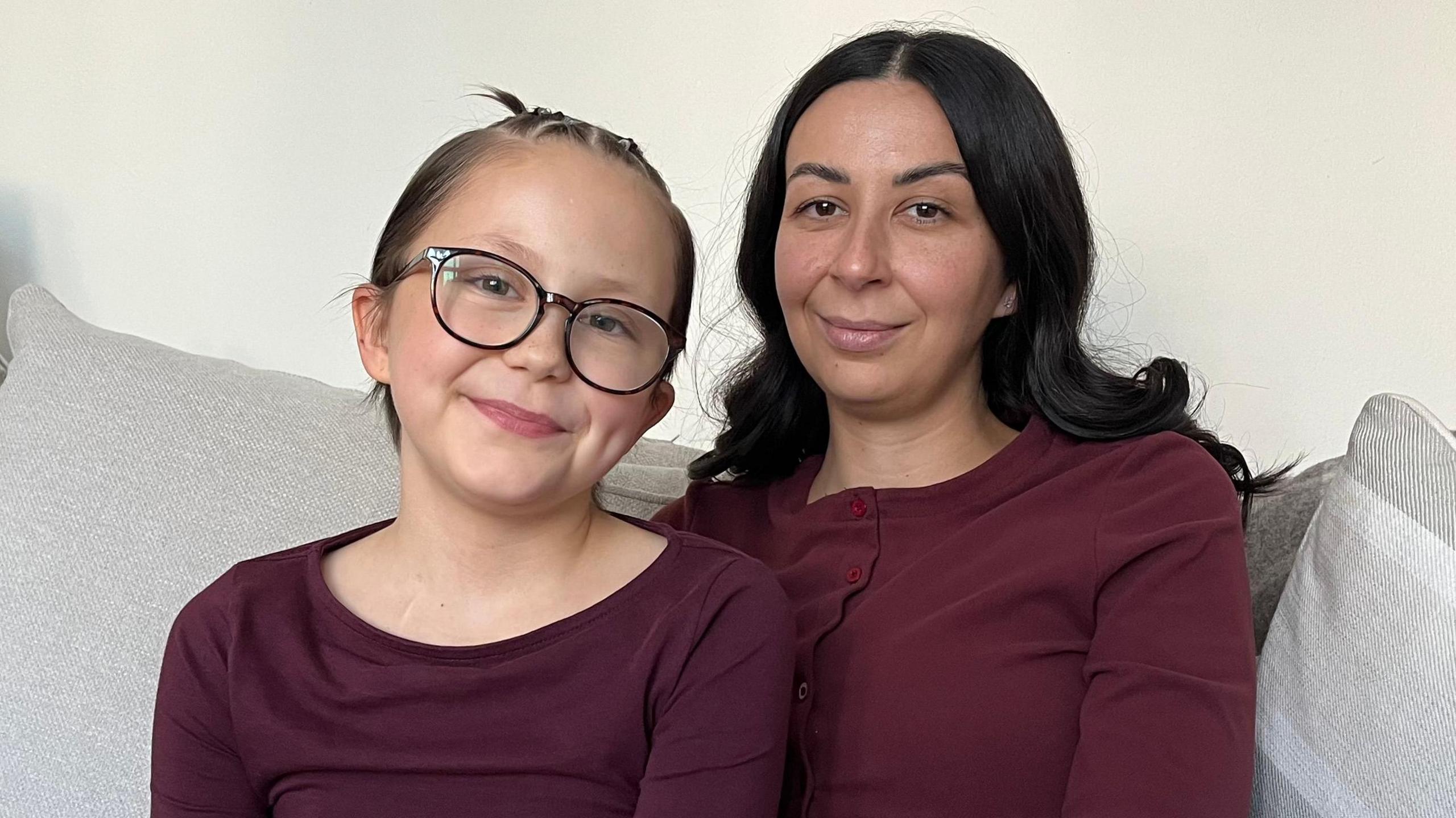 A woman in her 30s with dark long wavy hair, olive skin and brown eyes sits next to a girl of 10 with short brown hair which is braided on top, and she is wearing black framed glasses. They are sitting on a grey sofa both smiling at the camera  and are both dressed in burgandy coloured tops, the wall behind them is painted cream. 