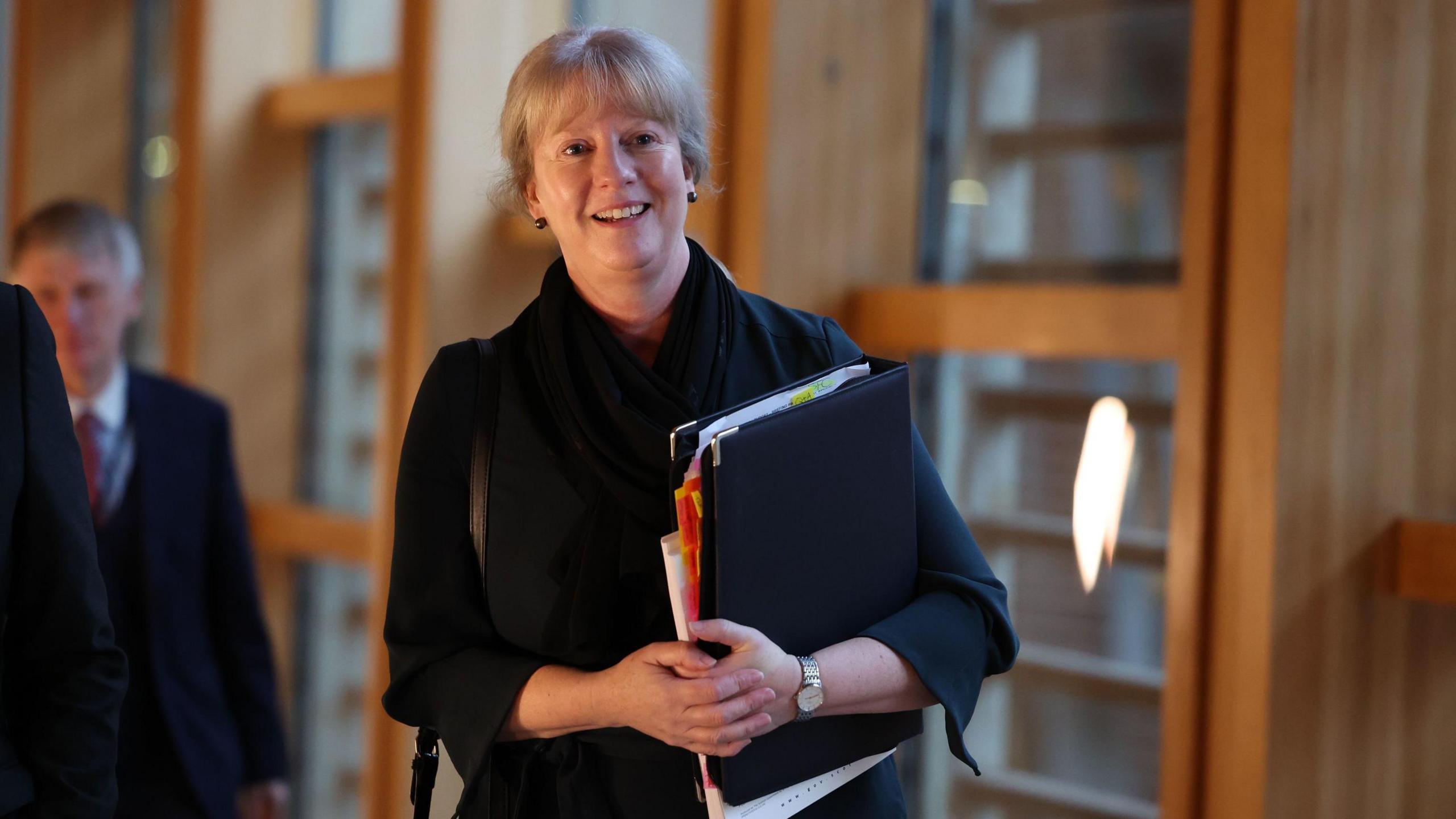 Shona Robison walking along the corridor at the Scottish Parliament. She has fair hair and is wearing a dark top. She is carrying a black folder, which has sections of paper coming out of the side. She is wearing a light-coloured watch on her left wrist.