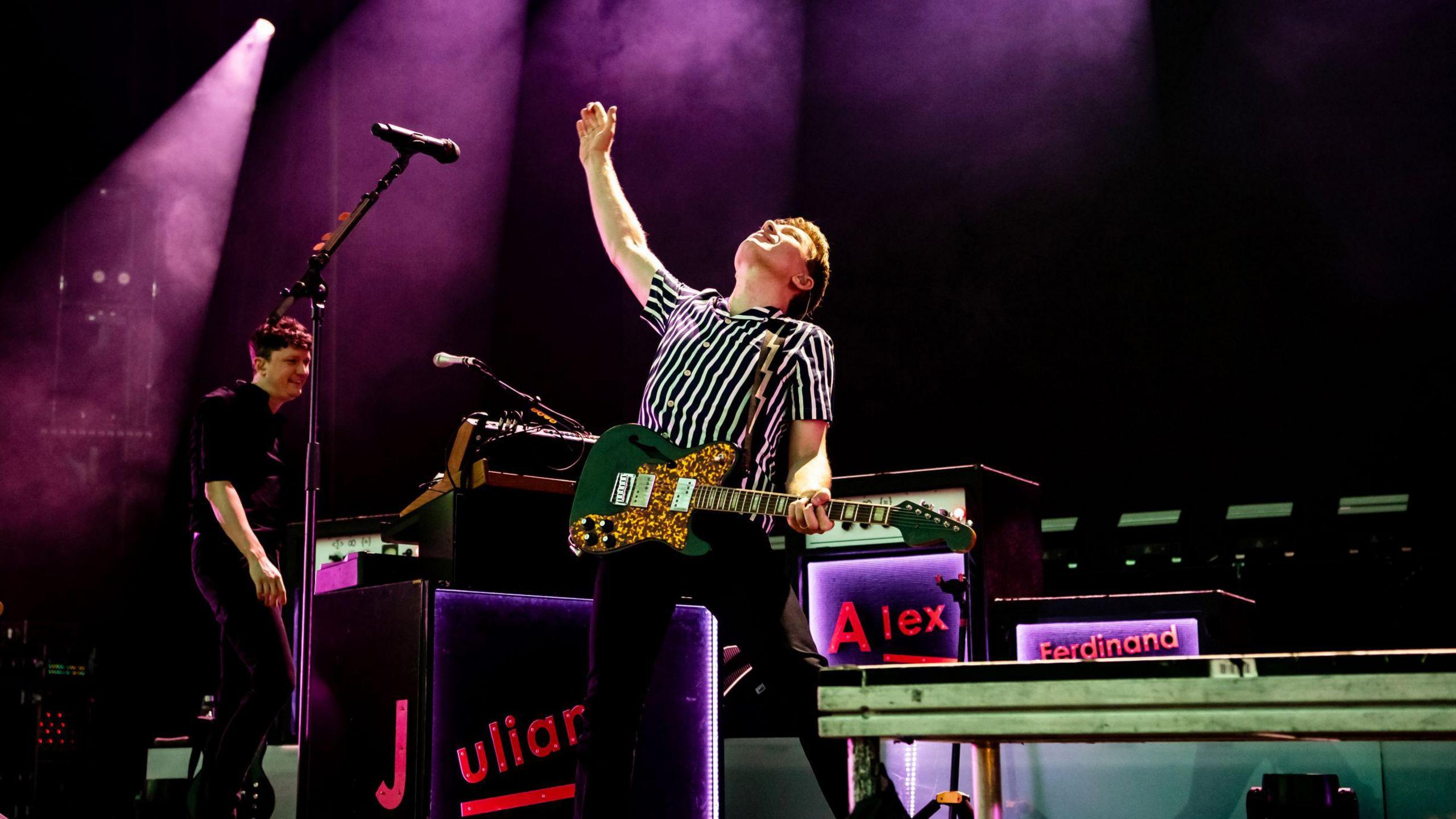 Alex Kapranos onstage at a Franz Ferdinand gig,  with one hand wrapped around his guitar and the other raised open-palmed in the air. Bandmate Julian Corrie can be seen playing guitar in the background, and the band's amps are visible too. 