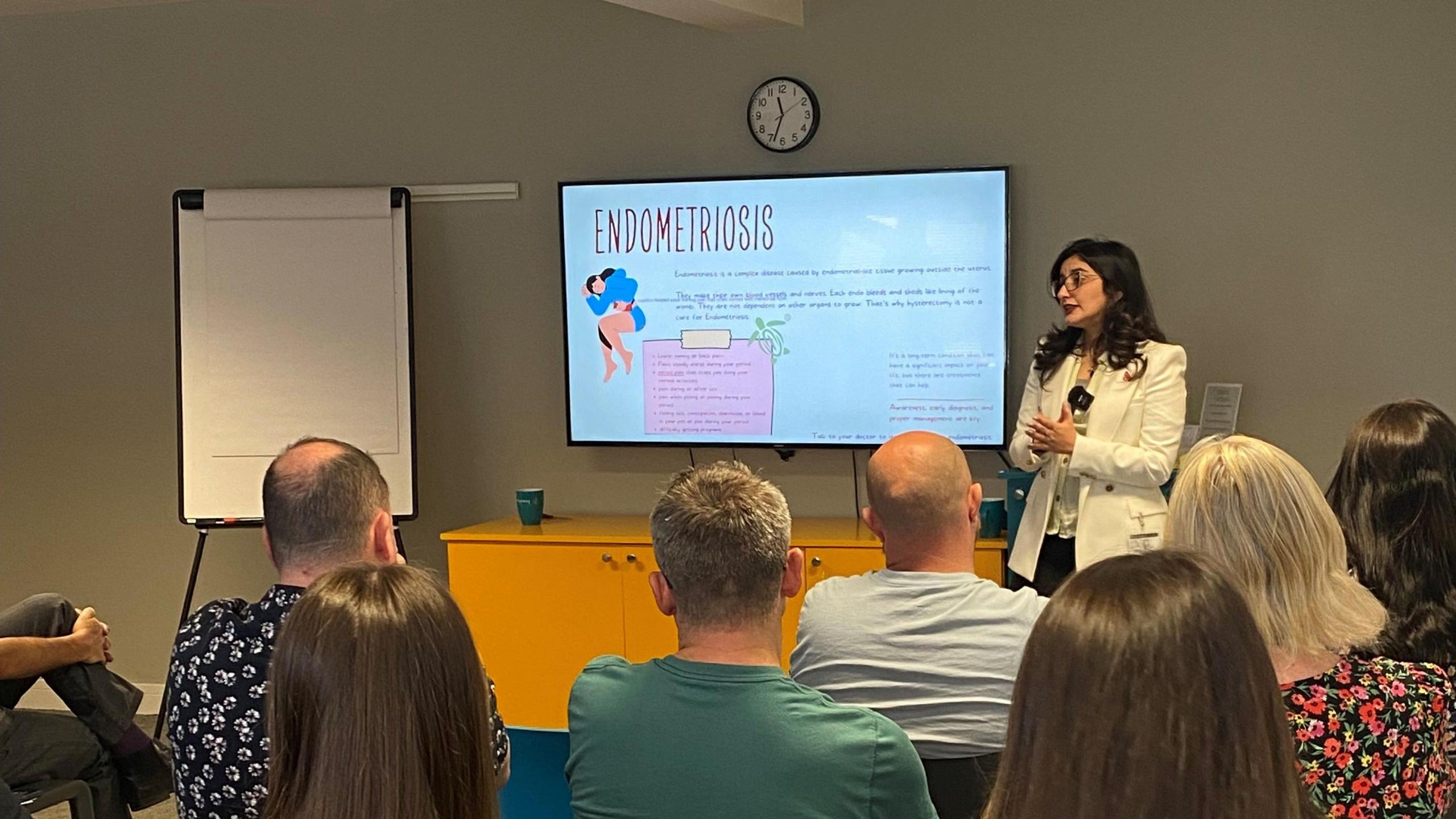 Roya Rasouli is standing in front of a screen which includes information about endometriosis. In front of her are number of people who are sitting down and listing to her talk about the conditon.