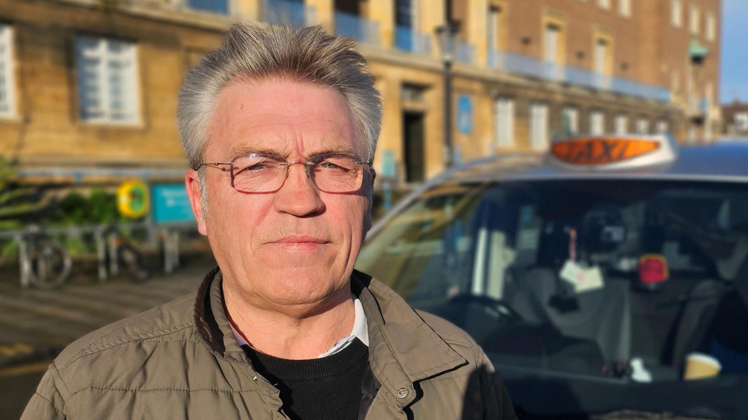 Craig Dimbleby standing outside Norwich City Hall, with his silver-coloured taxi behind him. He is wearing glasses and a green jacket over a black sweater and white shirt.