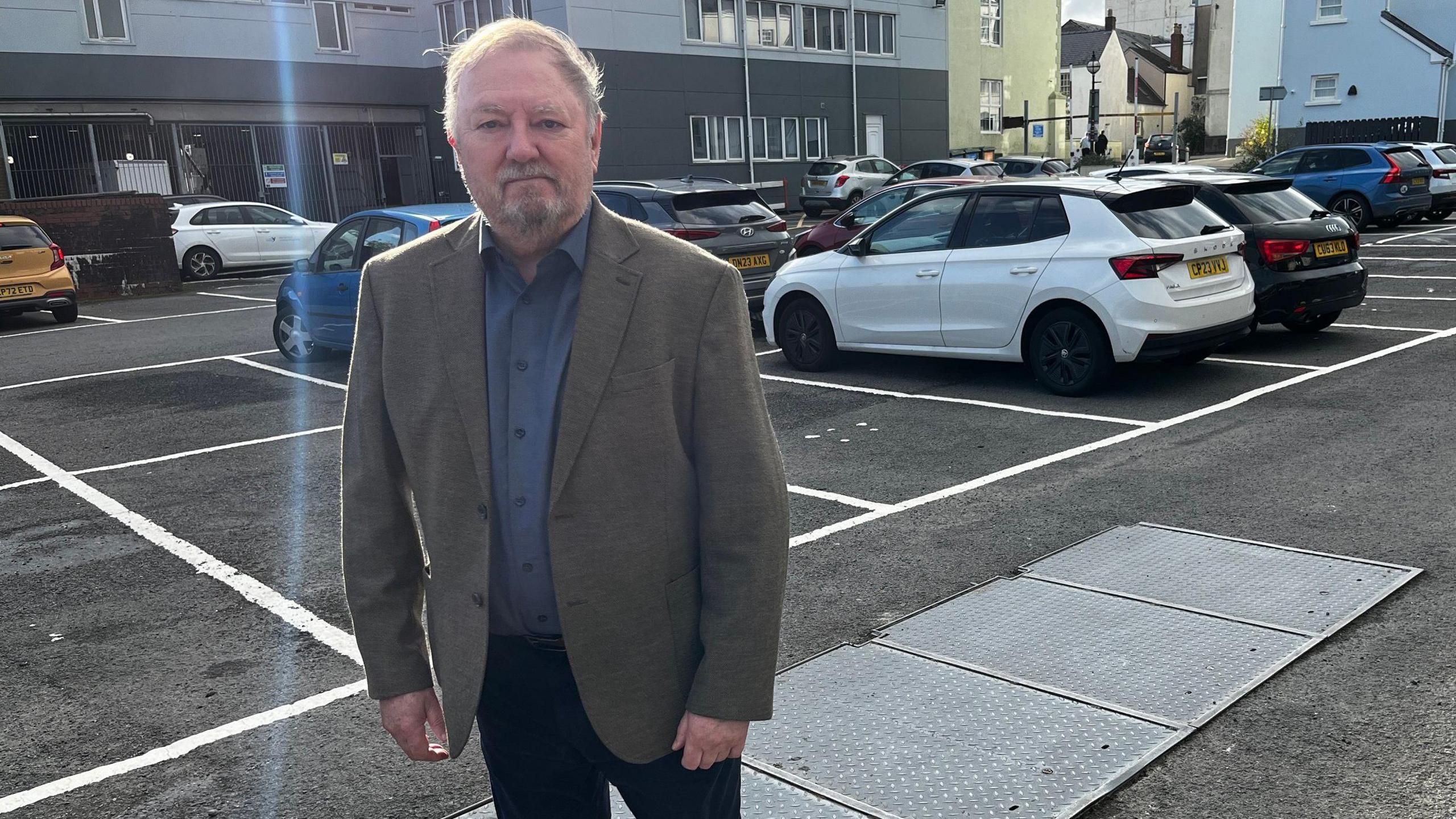 A man looks at the camera. He has a blue shirt and a grey jacket on with black pants. He has wispy grey hair and a grey goatee beard. The bunker can be seen bottom right of the image. As well there are cars that can be seen in the car park.