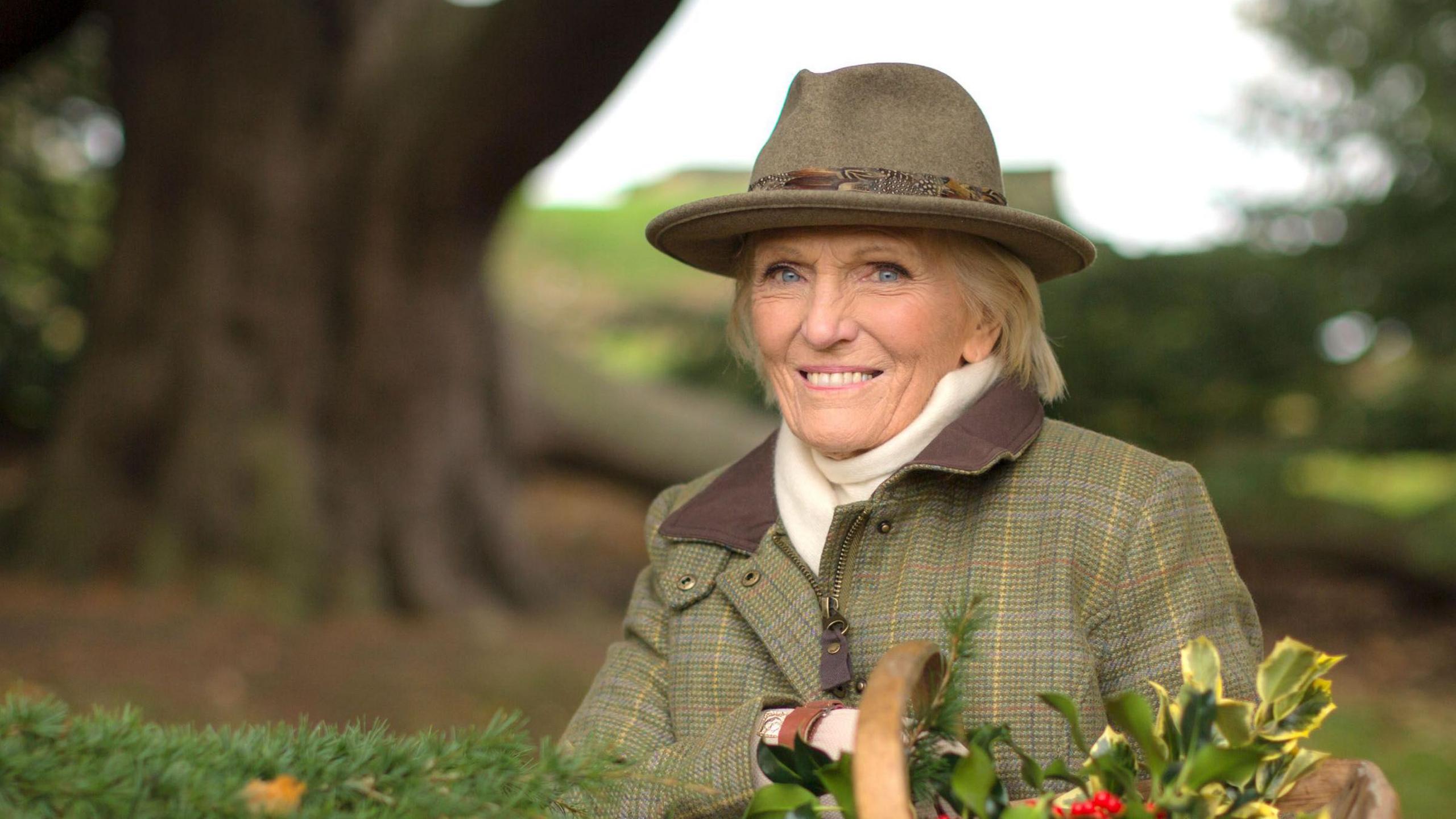 Dame Mary Berry holding holly in a basket and smiling at camera.