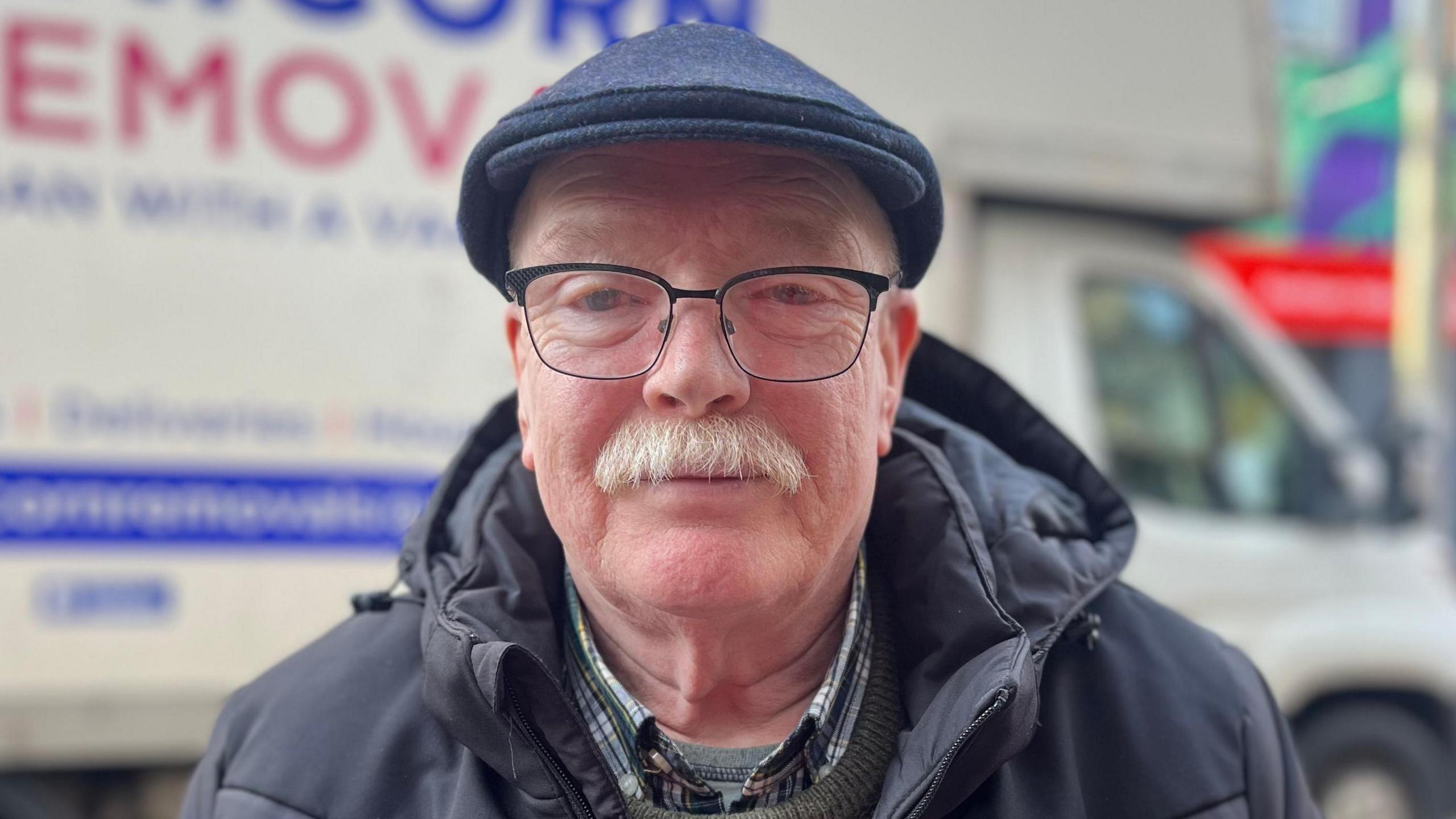 Mr Mitchell standing on the street in front of a white moving van. He is wearing a navy flat cap, metal glasses, checked shirt, grey jumper and black puffer coat. He has a grey moustache.