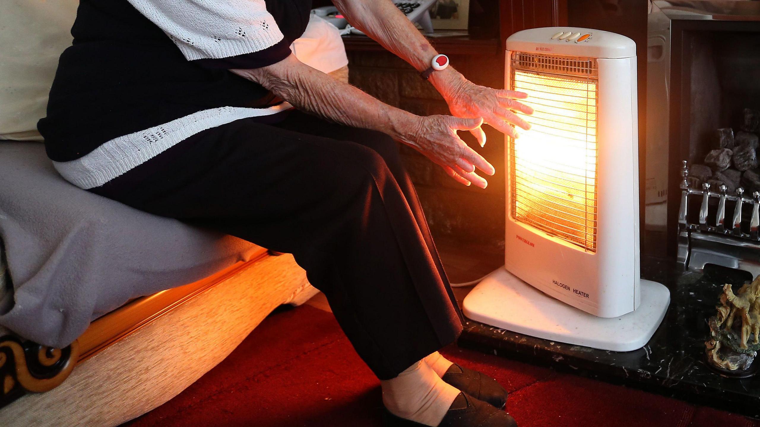 A pensioner, sitting down, holding their hands against a small electric heater