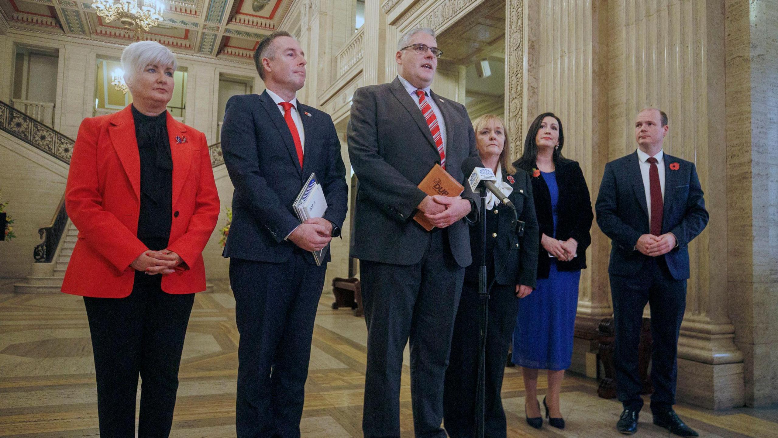 (Left-right) A DUP delegation including short blonde haired Joanne Bunting MLA in a red blazer, black trousers and top, Education Minister Paul Givan in a navy suit and red tie, party leader Gavin Robinson MP in a grey suit with red tie - both men have short grey hair - Michelle McIlveen MLA in a navy suit, with long blonde hair, Deputy First Minister Emma Little Pengelly in a blue dress, navy blazer, she has long brown hair and Communities Minister Gordon Lyons, balding in a navy suit and red tie, addressing reporters at Stormont on Monday. There are large marble stairs behind them