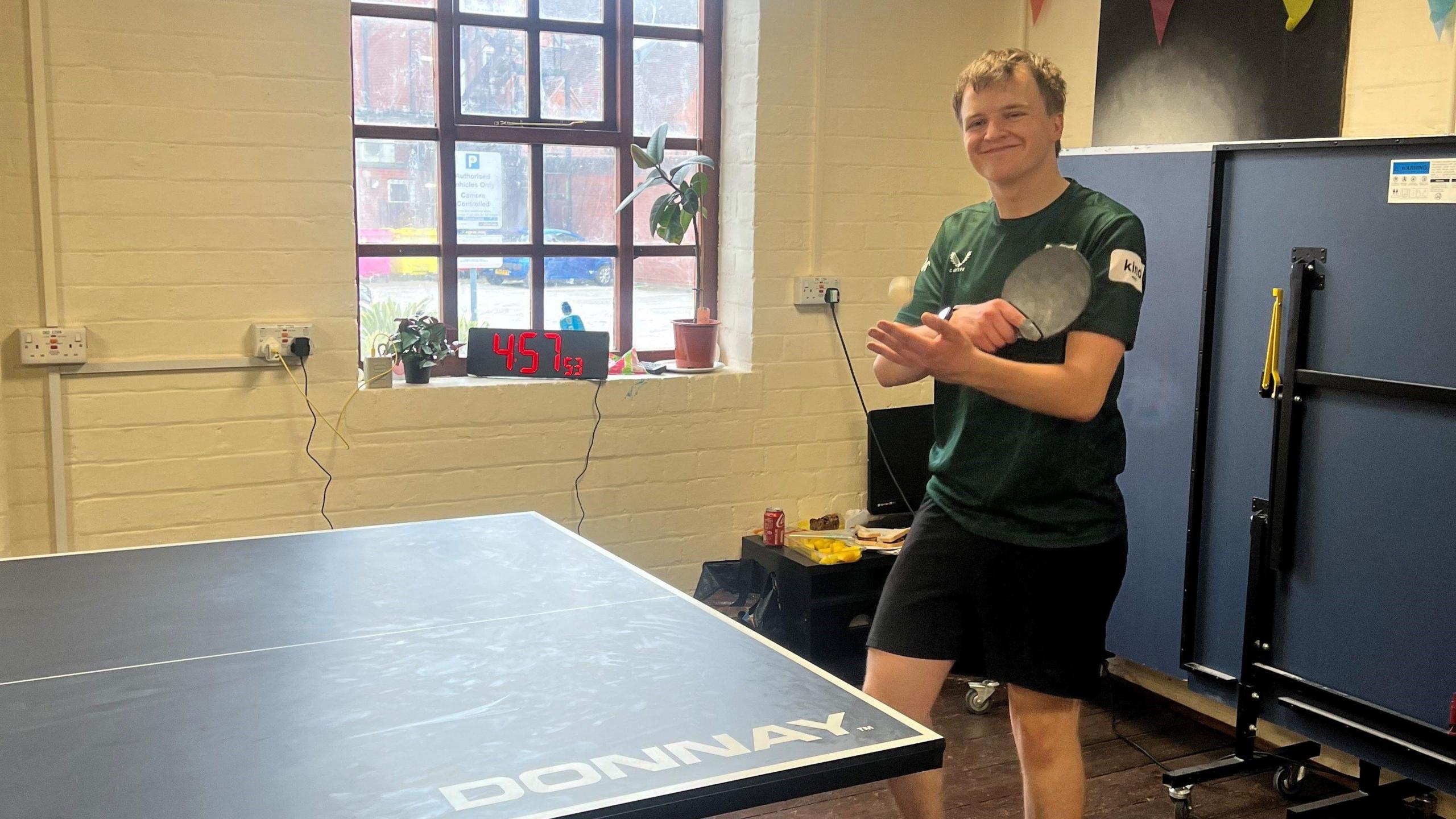 A young man with light brown hair wearing a dark green t-shirt and black shorts. He is holding a black table tennis bat and a white ball, in front of a blue table tennis table
