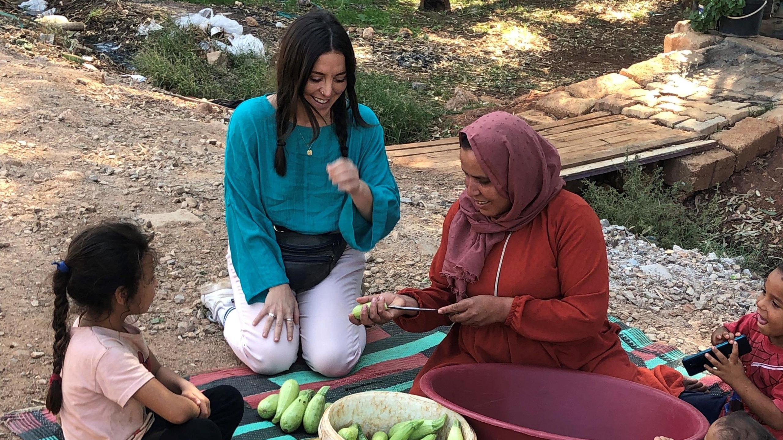 A young white woman with plaited black hair sits with a woman in a headscarf and two children on a blanket in a Syrian refugee camp