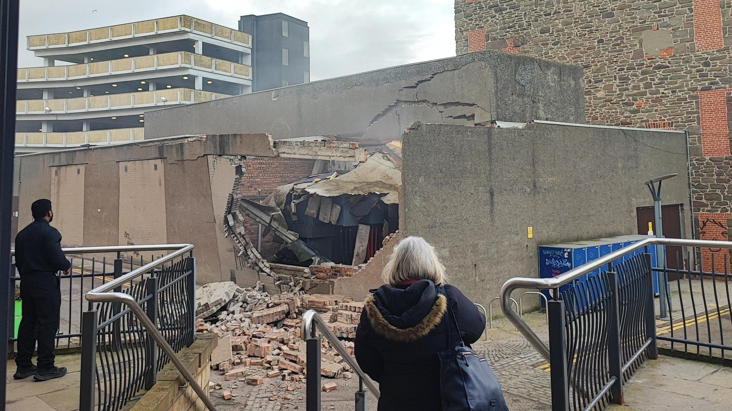 A badly damaged wall, with bricks strewn on the ground, following an explosion. Two bystanders survey the scene. 