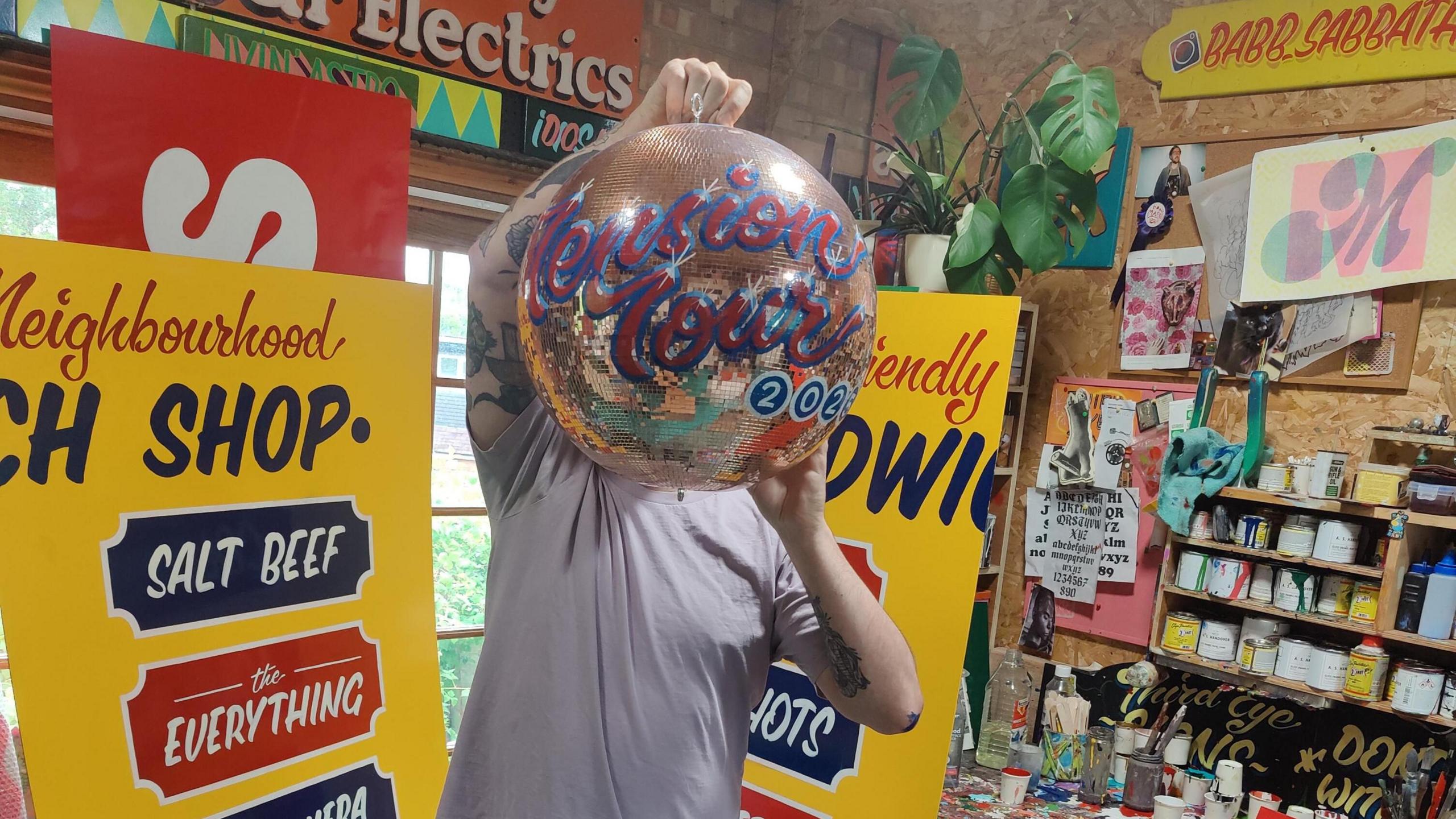 A man in a grey t-shirt holds up a mirrorball painted in red and blue letters which read 'Tension Tour' - he is in his workshop with painted signs all around. 