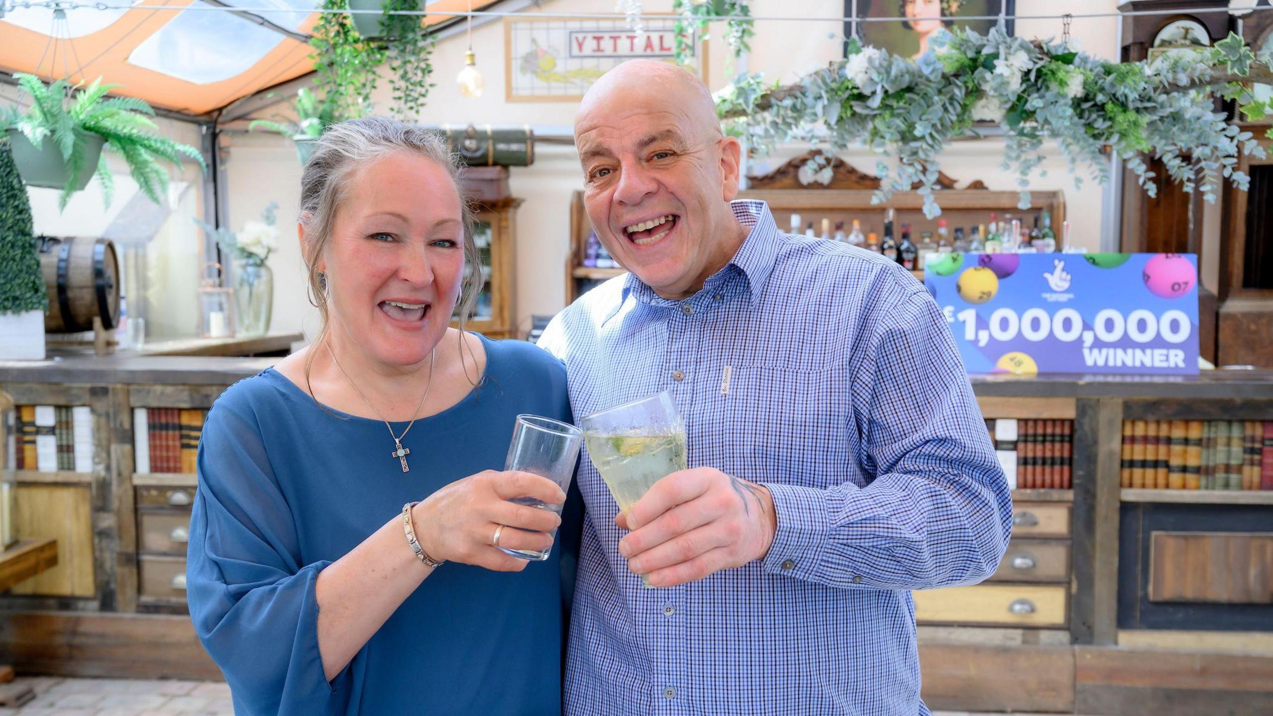 Caroline (left) and Terry Gillings celebrating their win with a drink. Caroline has grey hair, tied back, and is wearing a blue top. Her husband is bald and wearing a blue checked shirt. Behind them is a sign that reads "£1,000,000 winner".