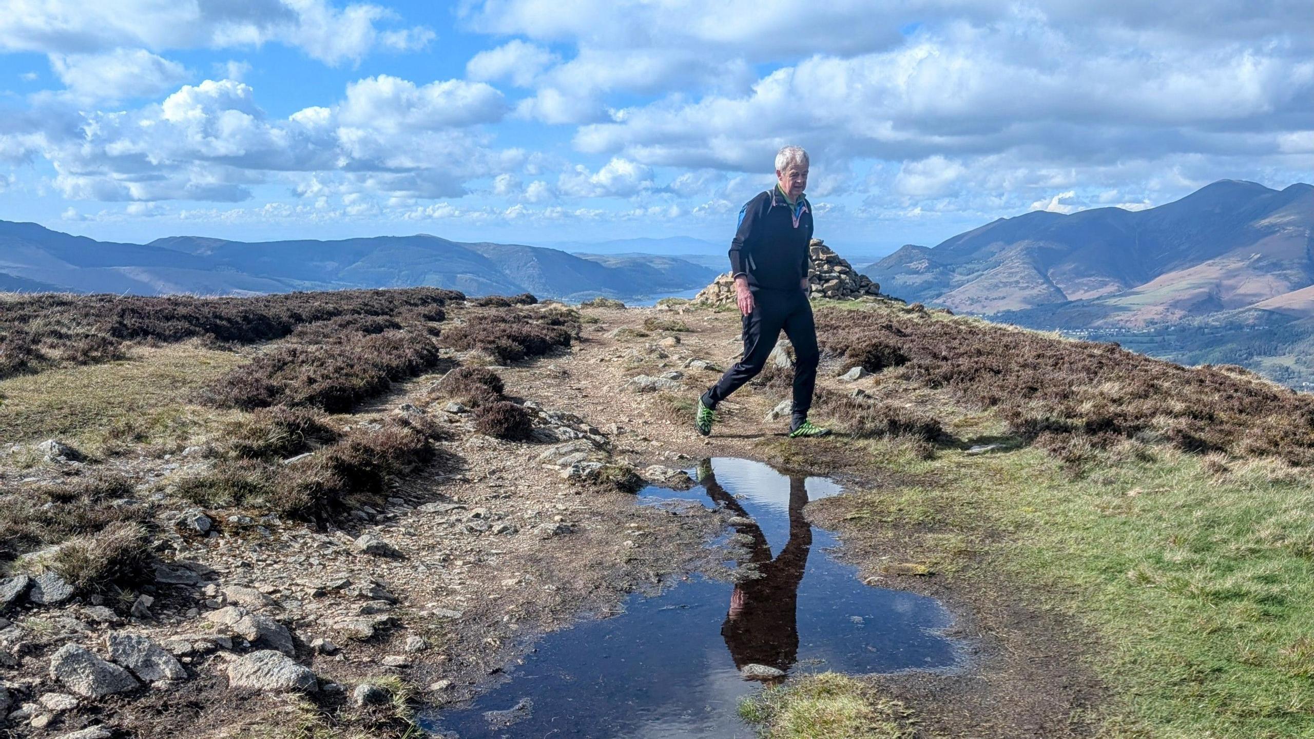 Ian on a hilltop walk. There's a rock pool of water reflecting his image. Behind him are mountain tops and hilltops. The sky is blue but cloudy. Ian is dressed in waterproof walking clothing including a black top and trousers. His green shoes are walking on the rocky ground beneath him.  