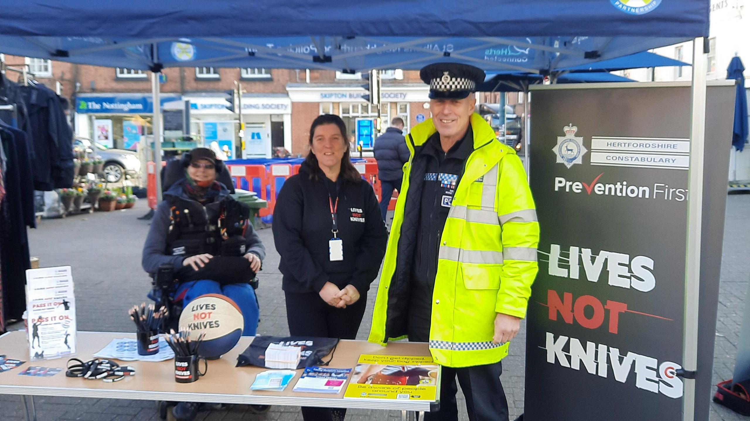 A stall run by Hertfordshire Constabulary at St Albans market. There three people looking straight at the camera, one officer, in full uniform with a high vis jacket. They have a table with pens, lanyards and leaflets on it. You can see market stalls behind and banner to the right. They are under a blue gazebo. 