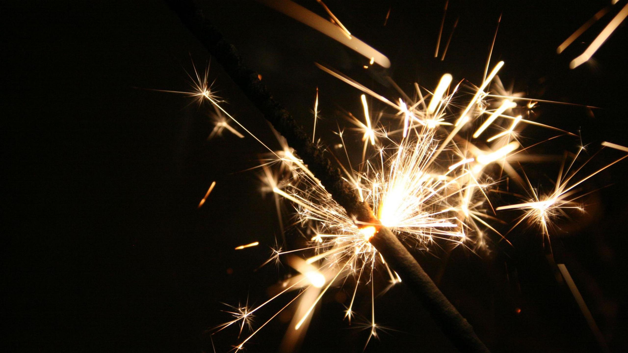 A close-up of a lit sparkler on bonfire night, with bright white sparks coming from a burning stick