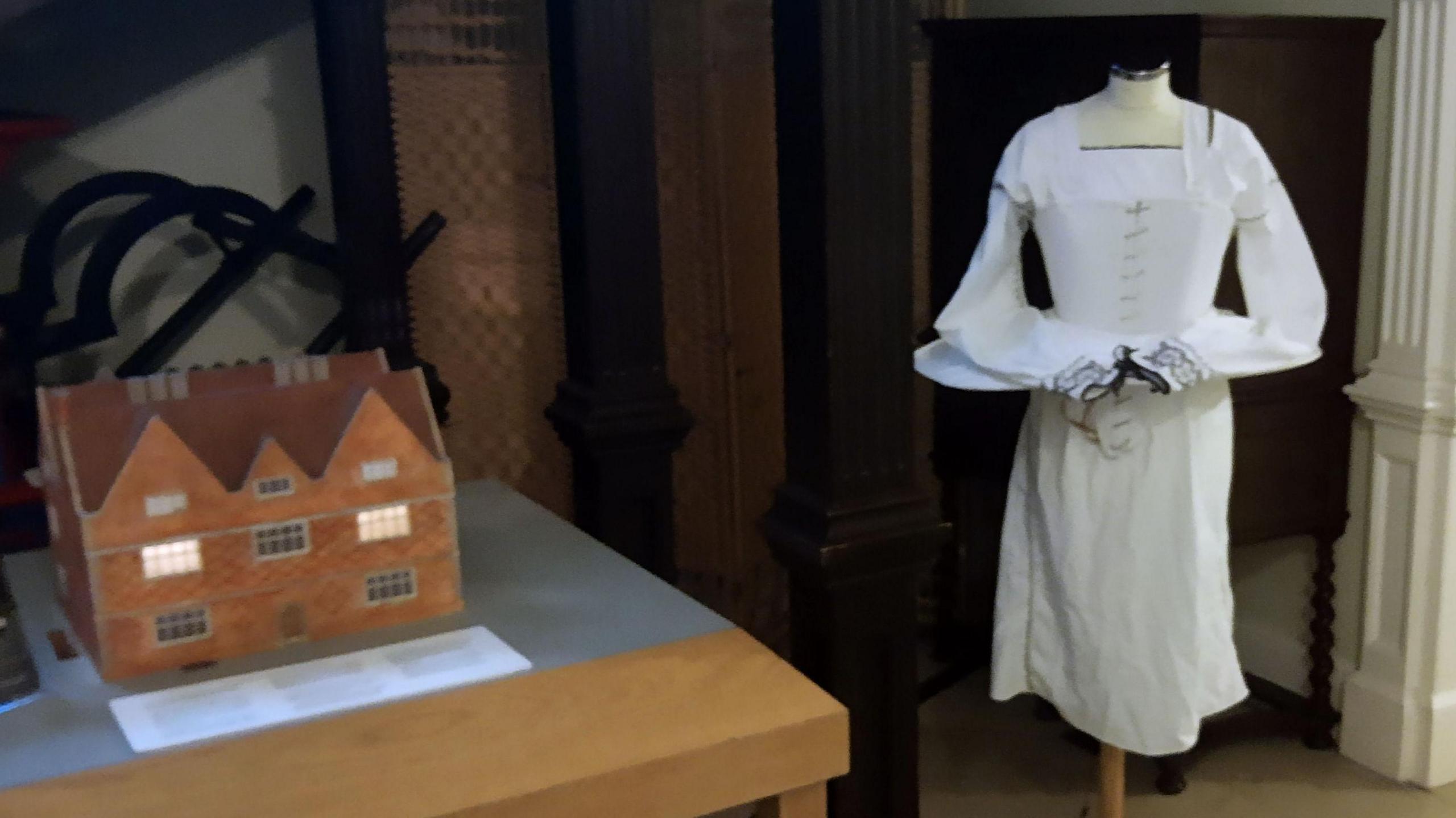 The image shows a museum exhibit with two displays. On the left, there is a scale model of a brick house with glowing windows, placed on a wooden table with an information plaque in front. On the right, a mannequin is dressed in a long white historical gown with lace sleeves. The background features dark wooden columns and panelling.