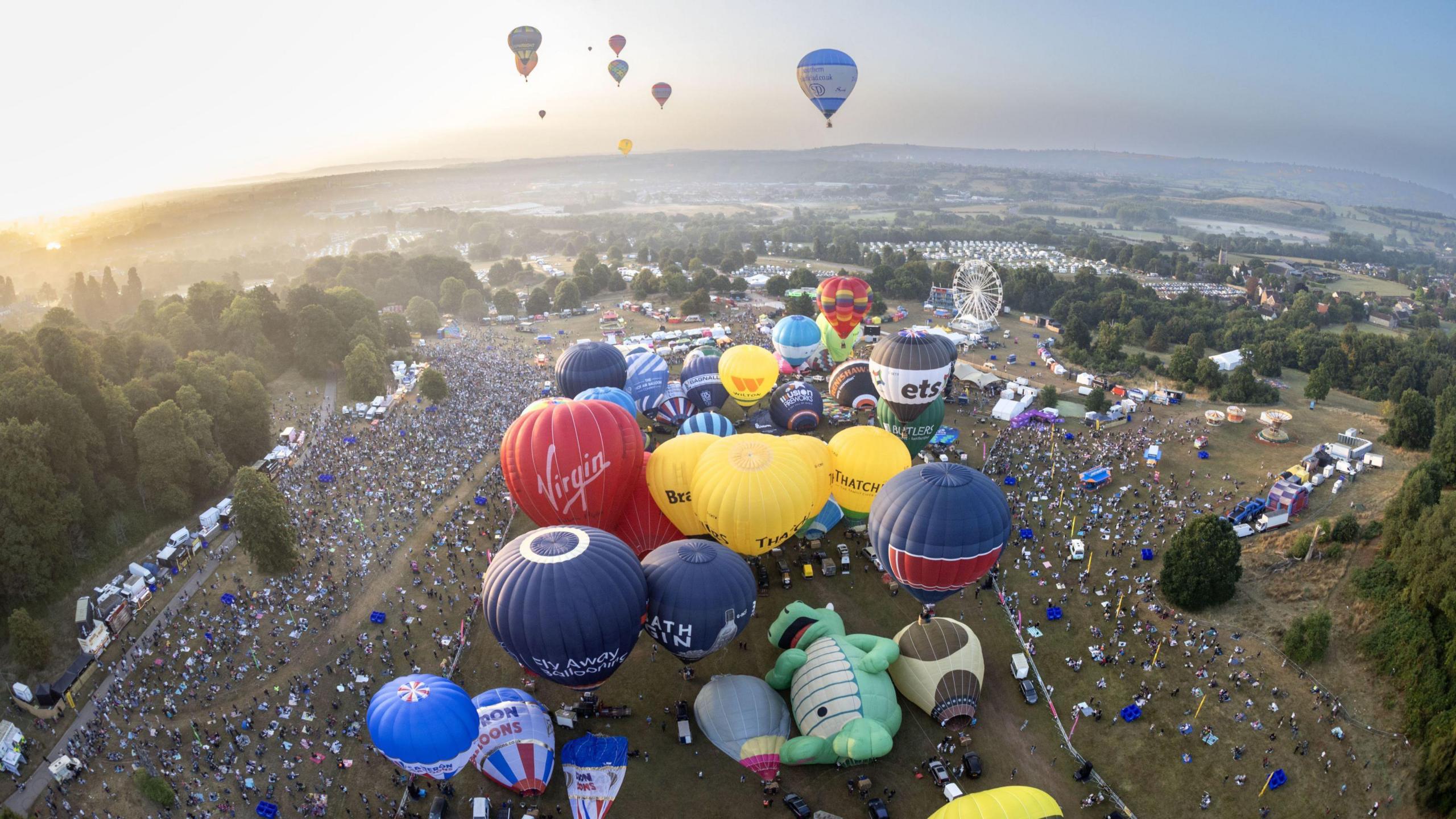 An aerial view of a dawn mass ascent at the Bristol Balloon Fiesta. The picture is taken from a balloon that has already taken off and is looking down at the main arena where dozens of other balloons are inflated ready to fly. They are many different colours and some have company branding on them. Around the edge of the launch field hundreds of spectators are visible and in the distance there is hazy light grey cloud on the hills on the edge of Bristol