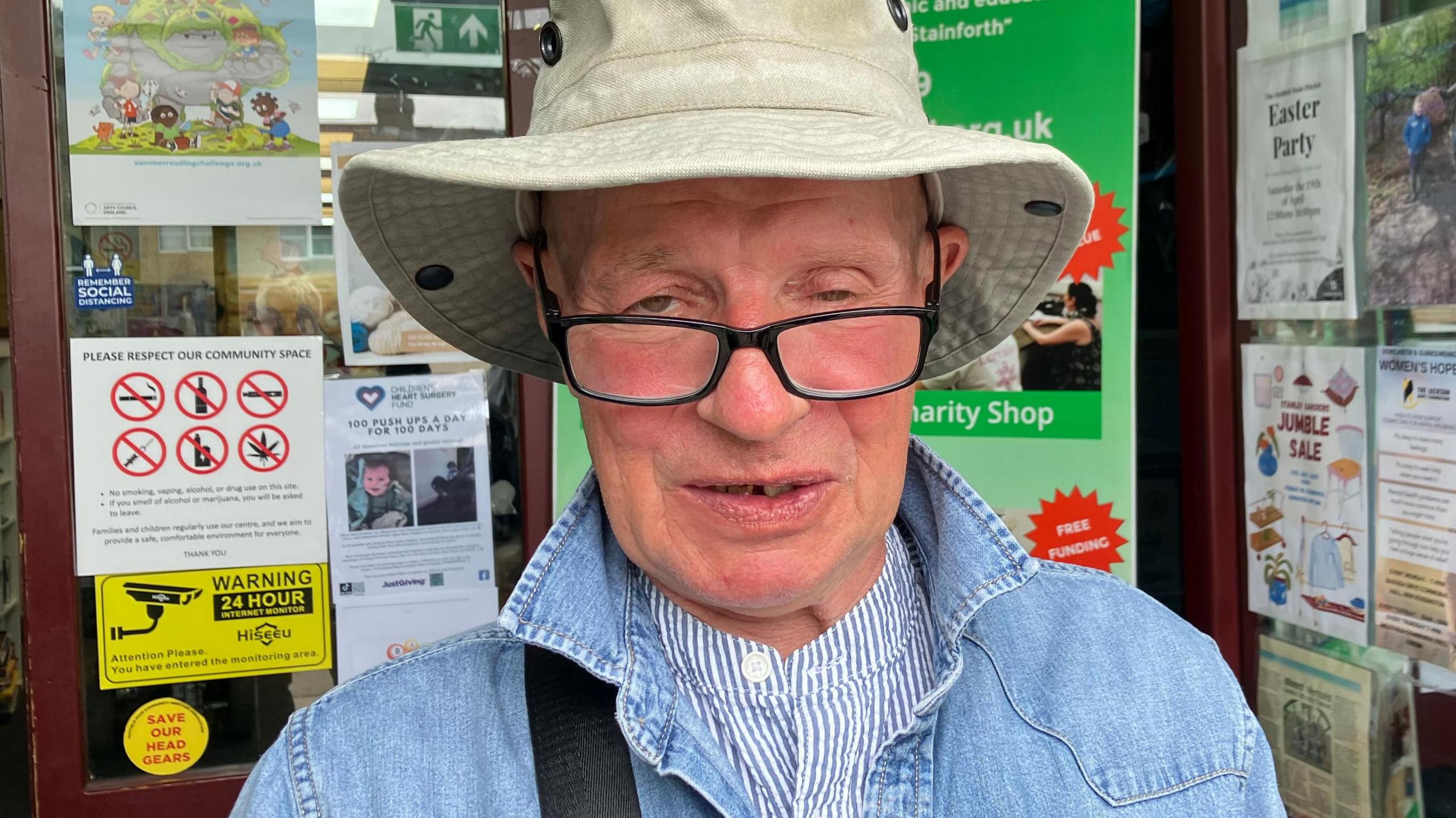 A man with black glasses and a fishing hat wears a blue jacket and a striped top. He is standing in front of a doorway.