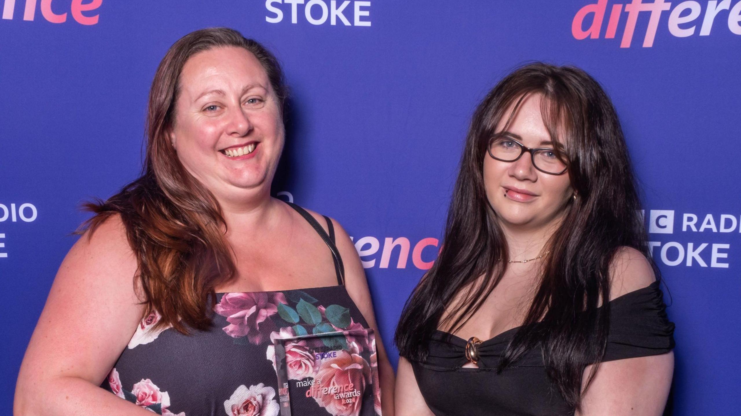 Two woman face the camera, smiling - one is wearing a floral patterned dress and the other is wearing a black dress