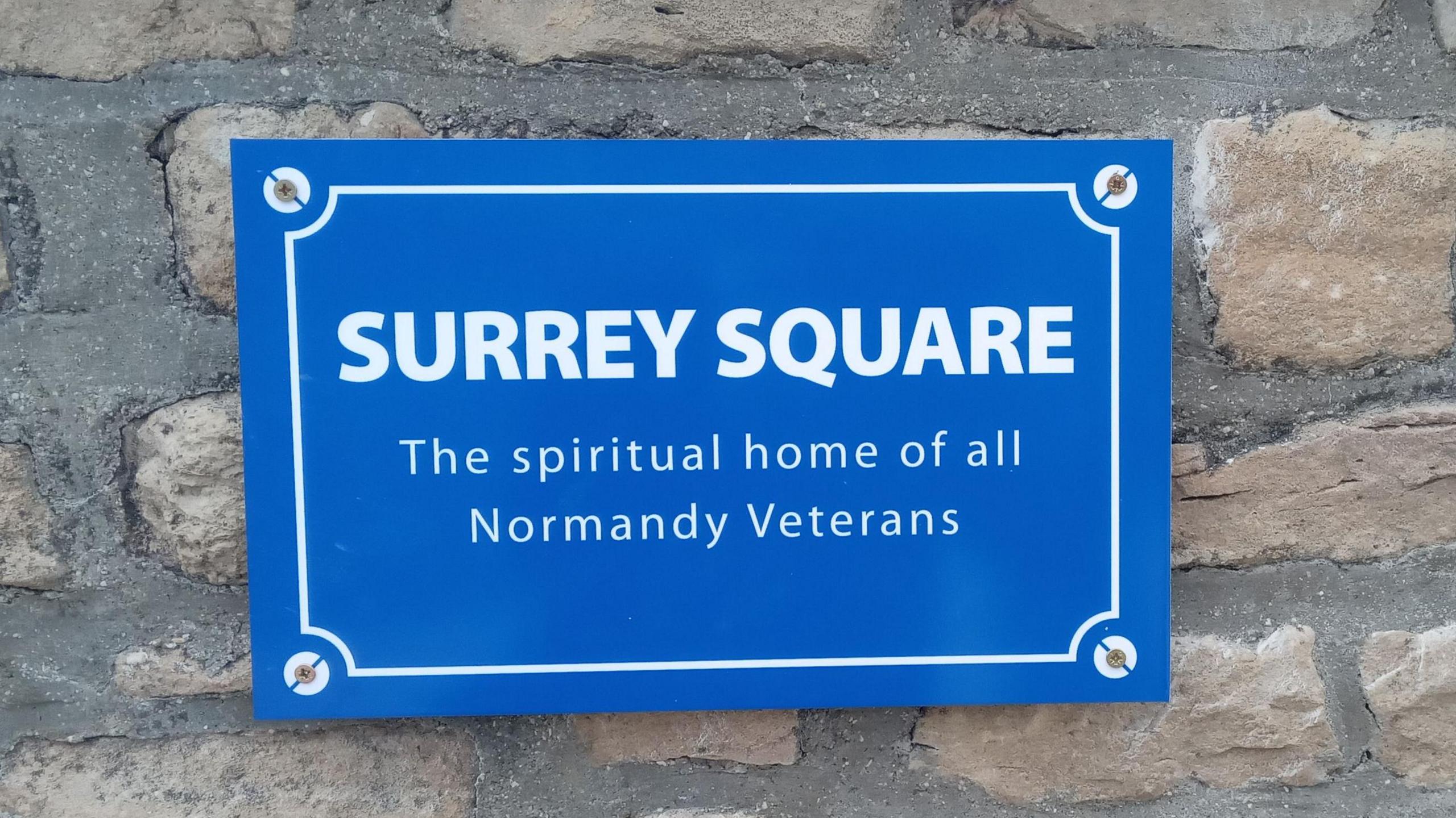 A blue plaque with white writing on a brick wall. The plaque reads: "SURREY SQUARE - The spiritual home of all Normandy Veterans".