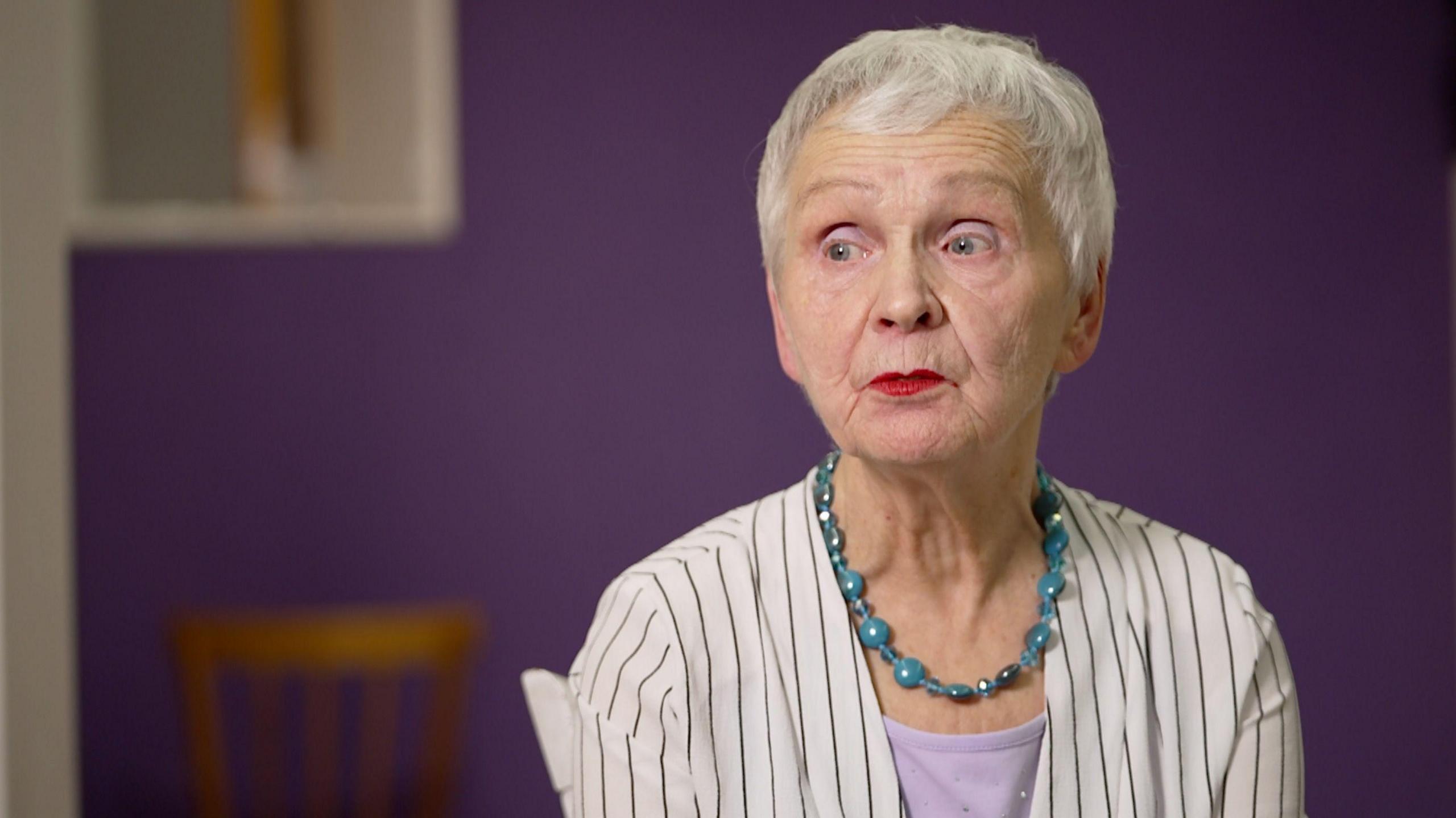 Ludmila Vasilyeva, 84, sits in a black and white vertical striped shirt, a turquoise bead necklace and pink shirt, facing camera left in front of a purple wall