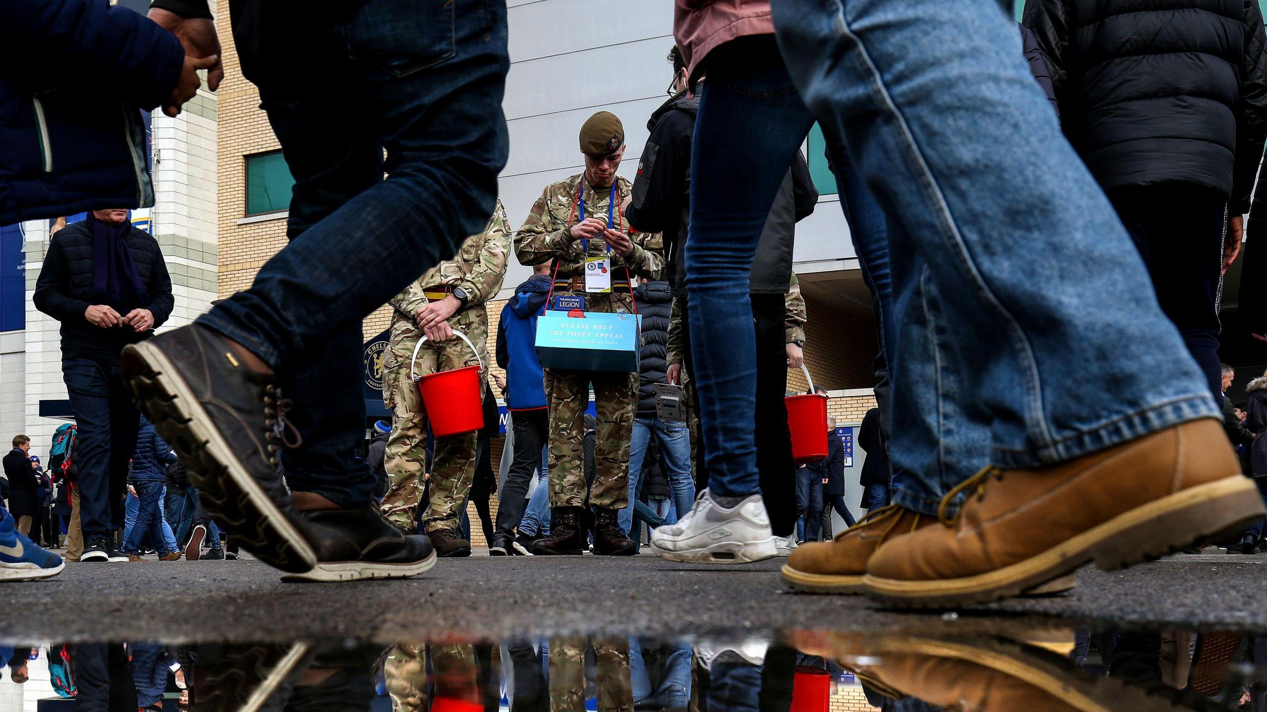 Armed Forces soldiers selling poppies in front of group of men who are stood near puddle.