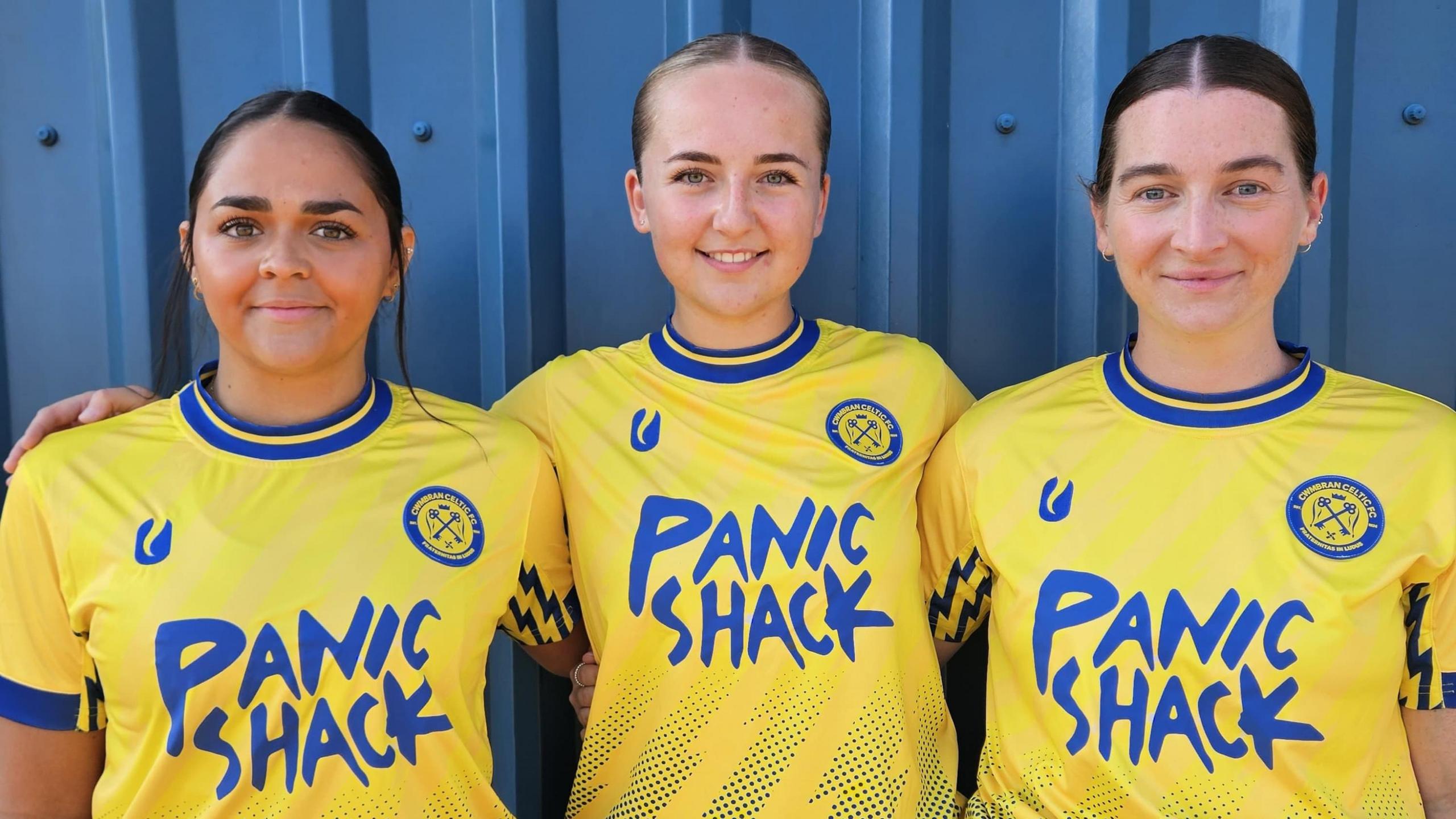 Three of the women's team players stand smiling at the camera in yellow home kit with blue sleeves and writing on the front