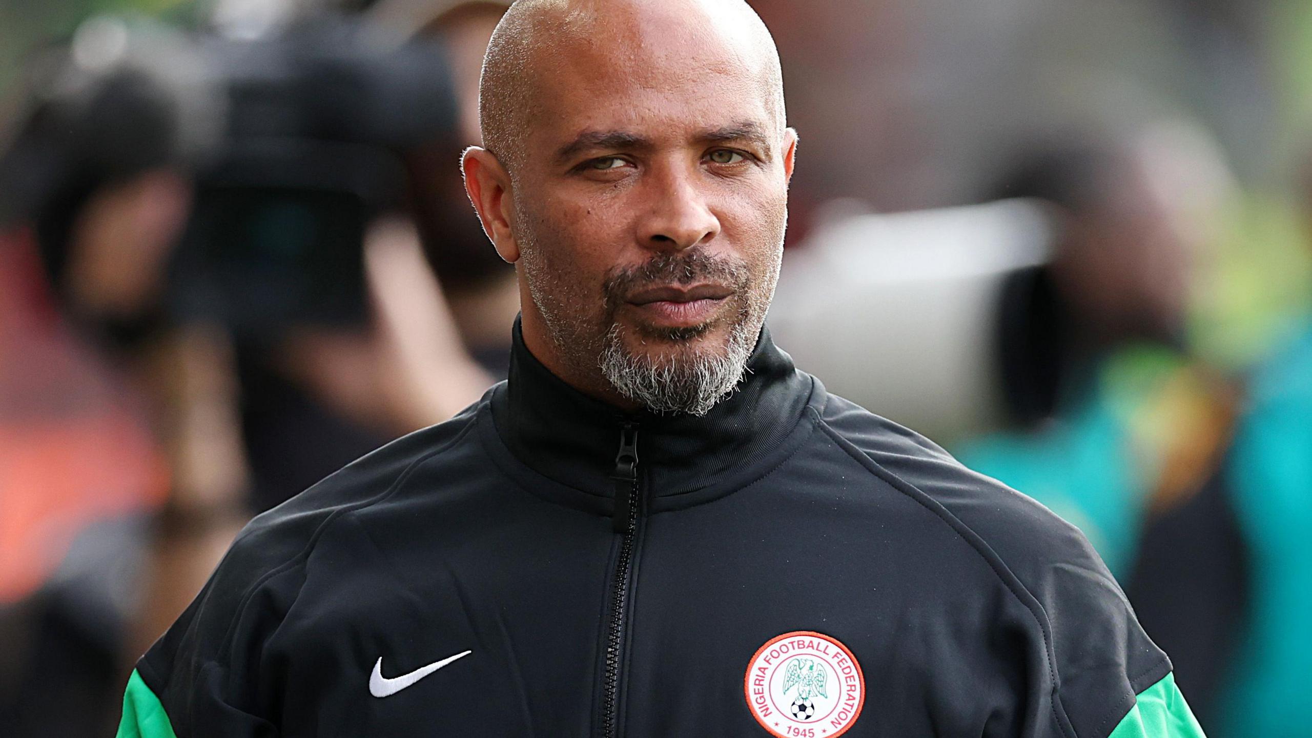 Eric Chelle, seen from chest up wearing a predominantly black tracksuit with green trim on the arms and a Nigeria Football Federation logo on the left breast, stands with a pensive look on his face. Chelle has a goatee beard flecked with grey and a shaved head