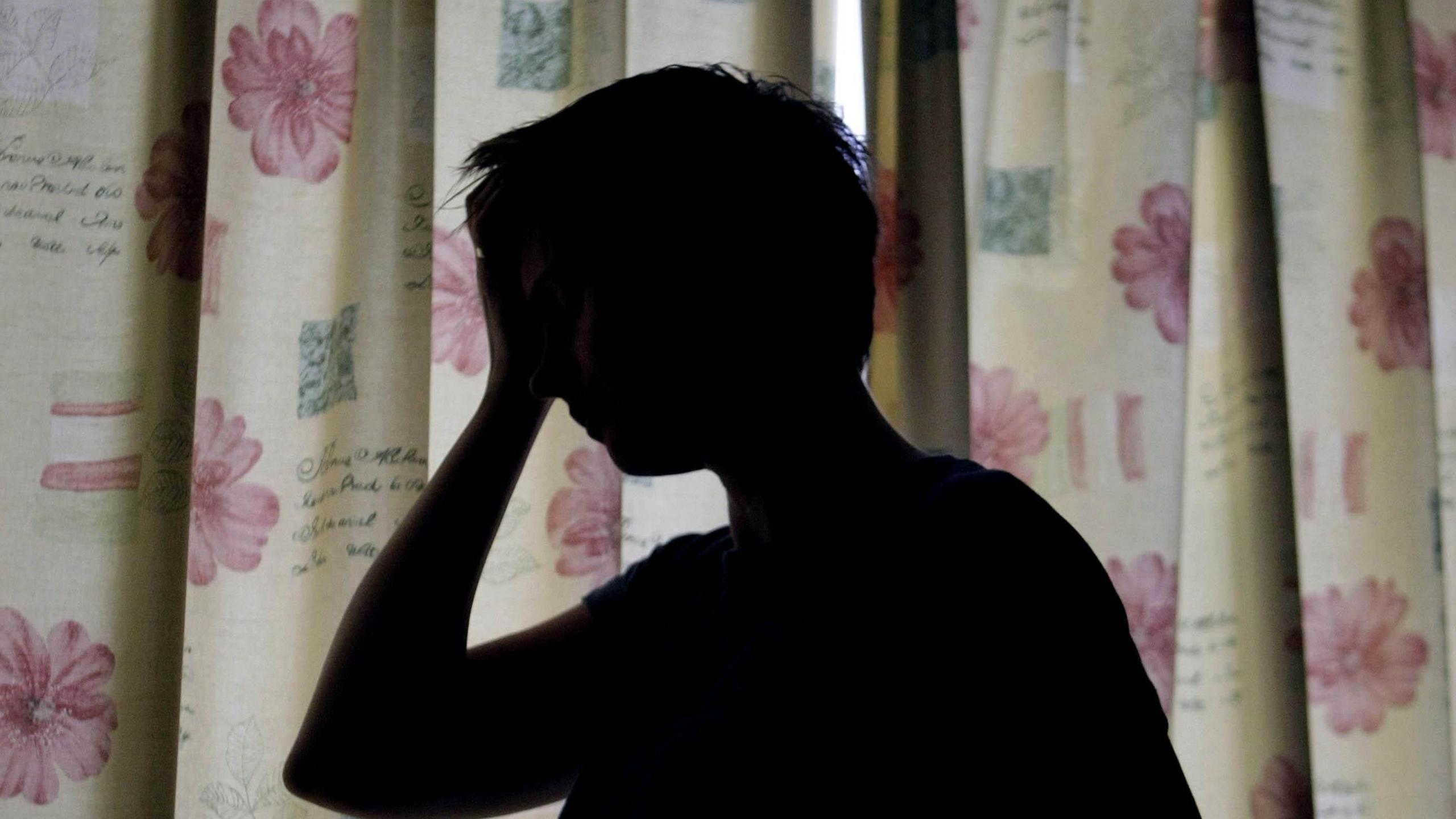 A generic image of a woman holding a hand to her head. She is blacked out while standing in front of yellow coloured curtains that have red flowers printed on them.