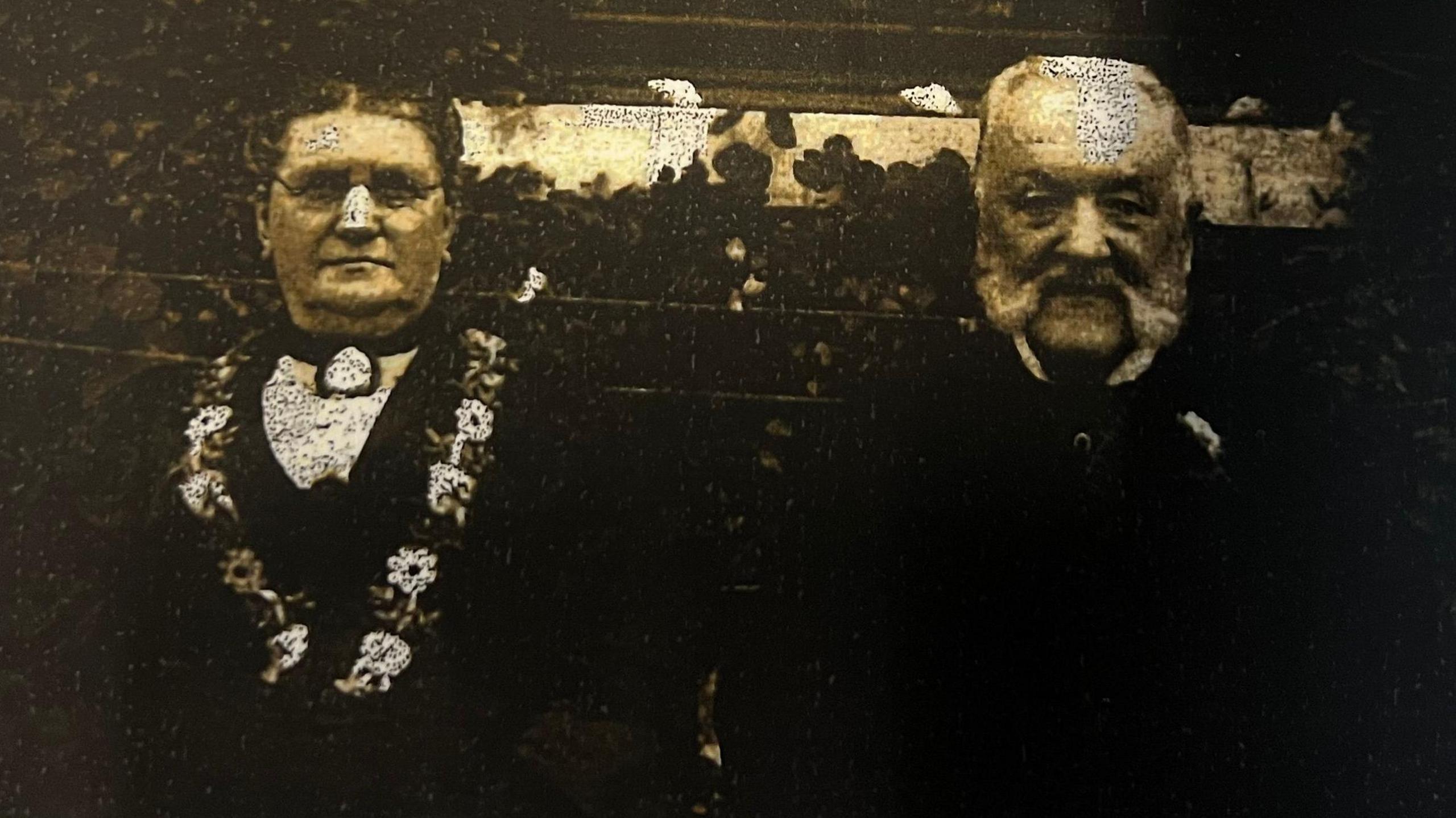 A man and woman, of late middle age, dressed in Victorian clothing. The black and white picture is very faded.