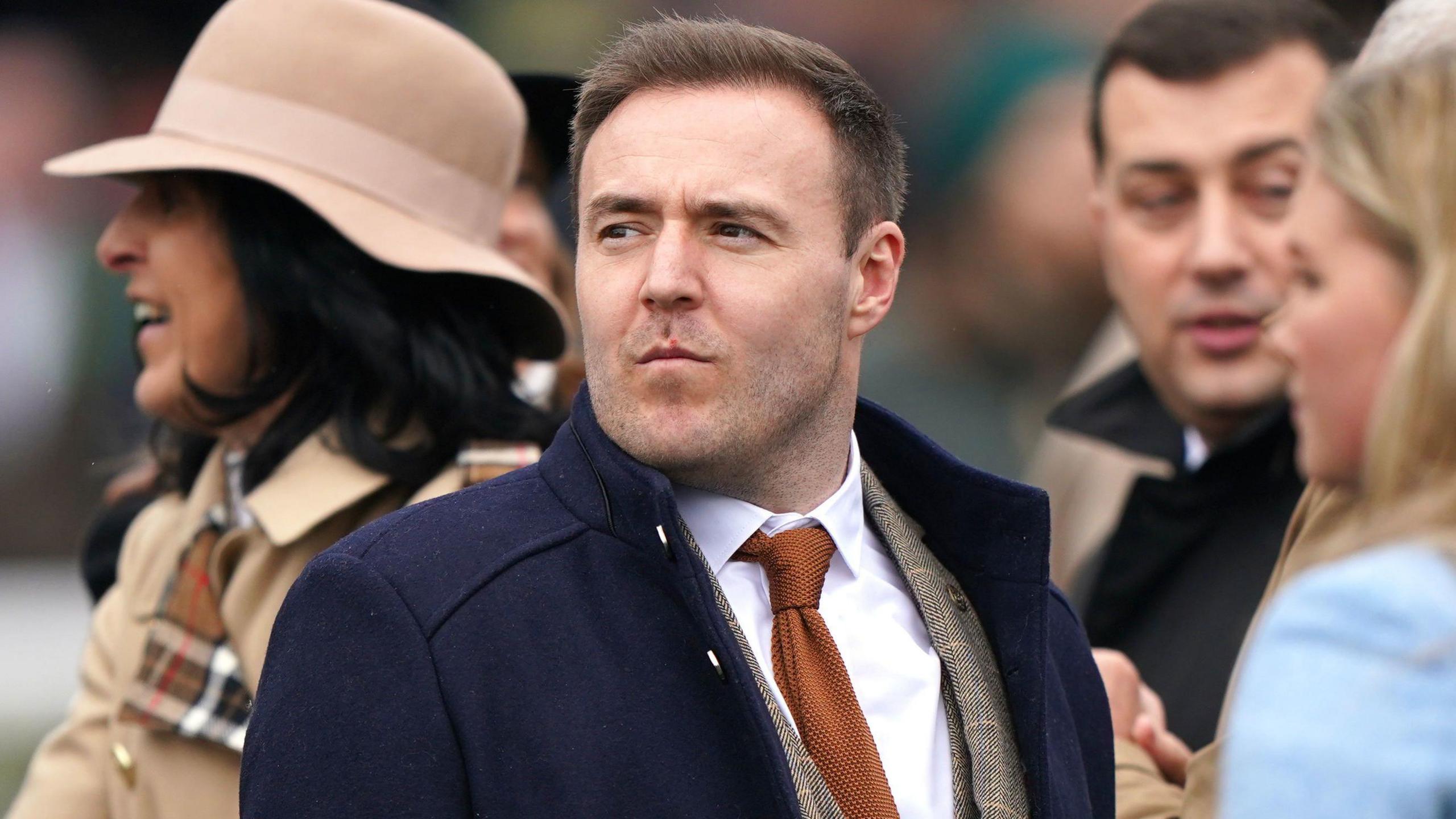 Alan Halsall in a tweed suit and black coat. He is amongst other people, whose faces are slightly blurred.