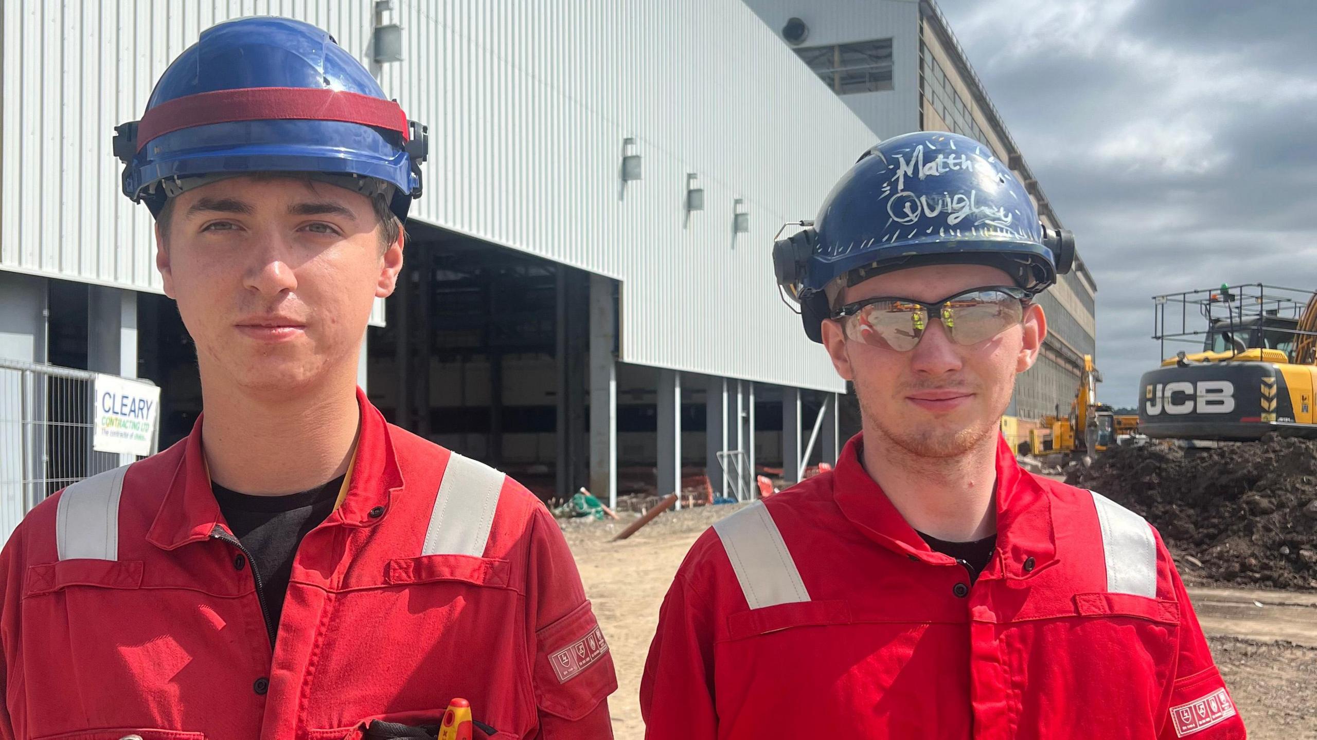 Keelan McCorriston and Matthew Quigley stand side by side in red work jumpsuits and blue hard hats. They are both young men and are smiling at the camera as they stand in front of the large metal warehouse.