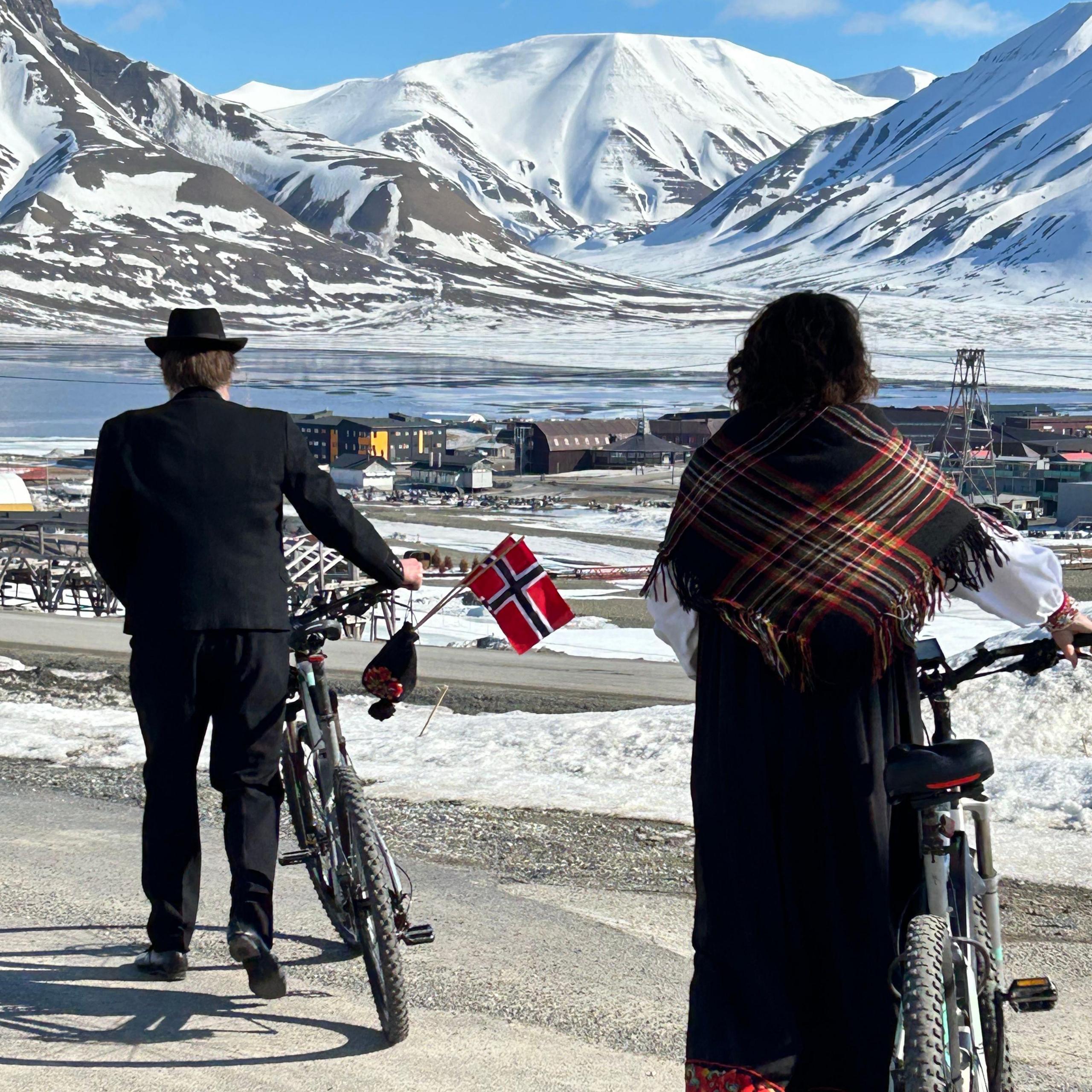 Norwegians wheeling their bikes on their national day in Svalbard, with water and icy peaks in the distance