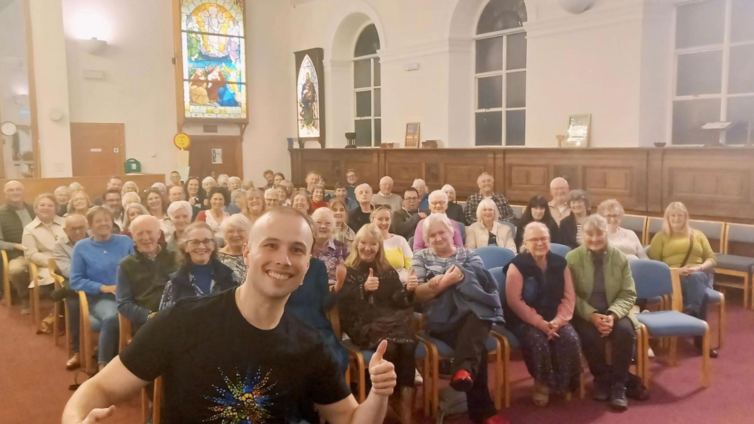 A smiling young man with a shaved head, and wearing a black t-shirt, puts his thumbs up as he stands in front of of a congregation of churchgoers.