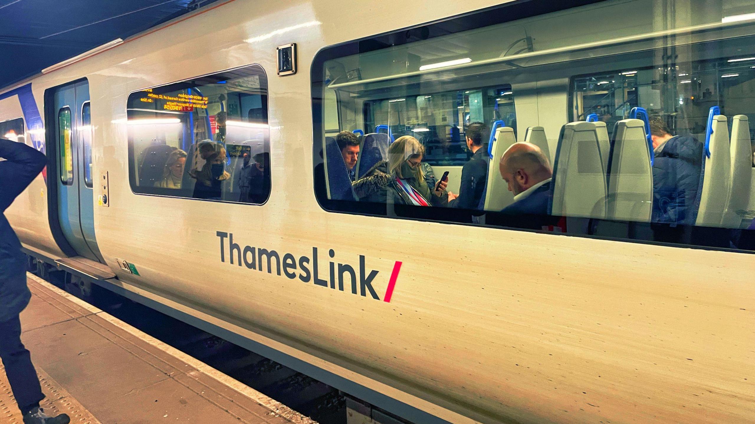A blue and white Govia Thameslink train at a platform, with passengers visible inside the carriage.