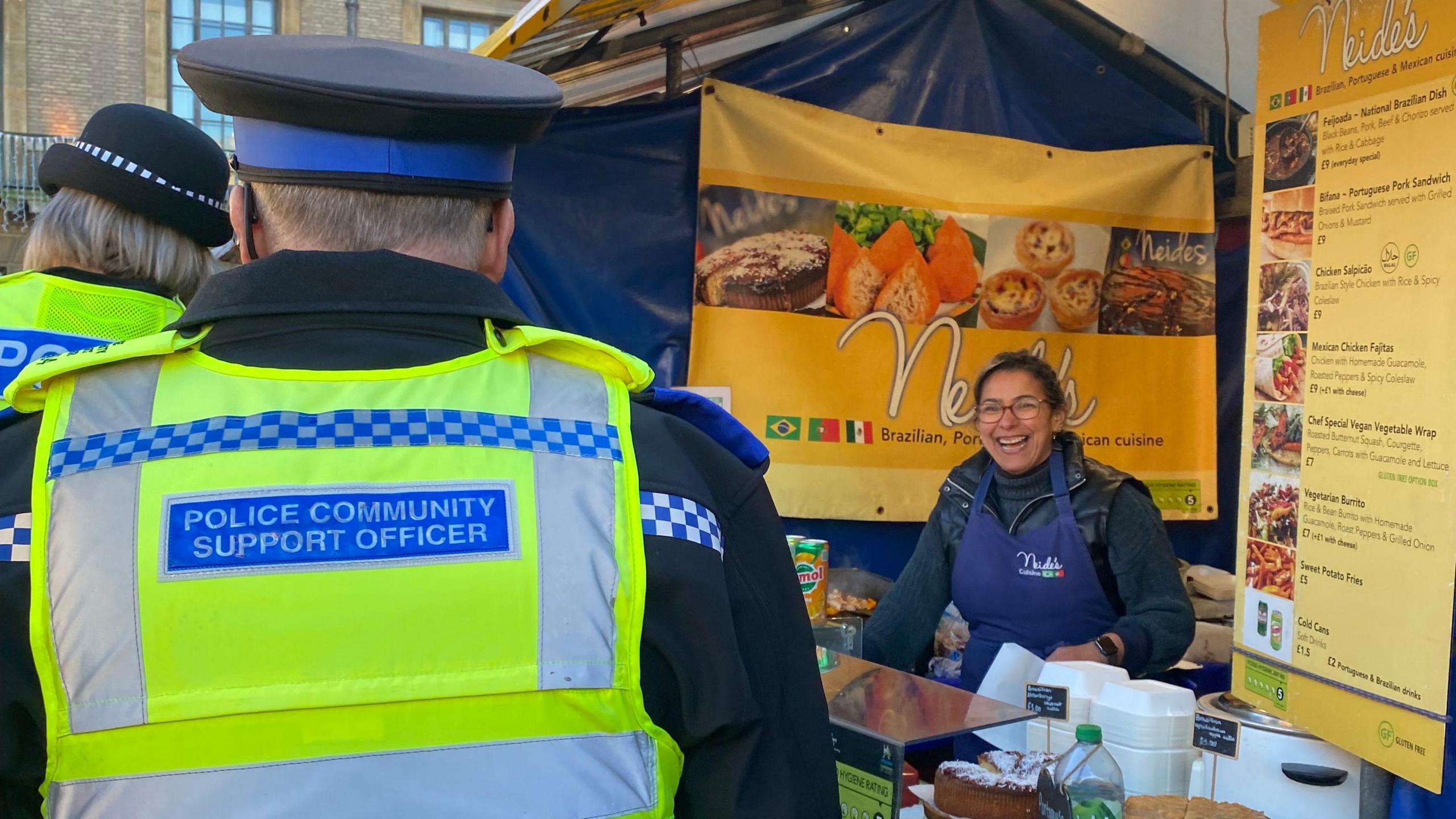 A market trader inside her stall wearing a blue apron smiling at two police officers 