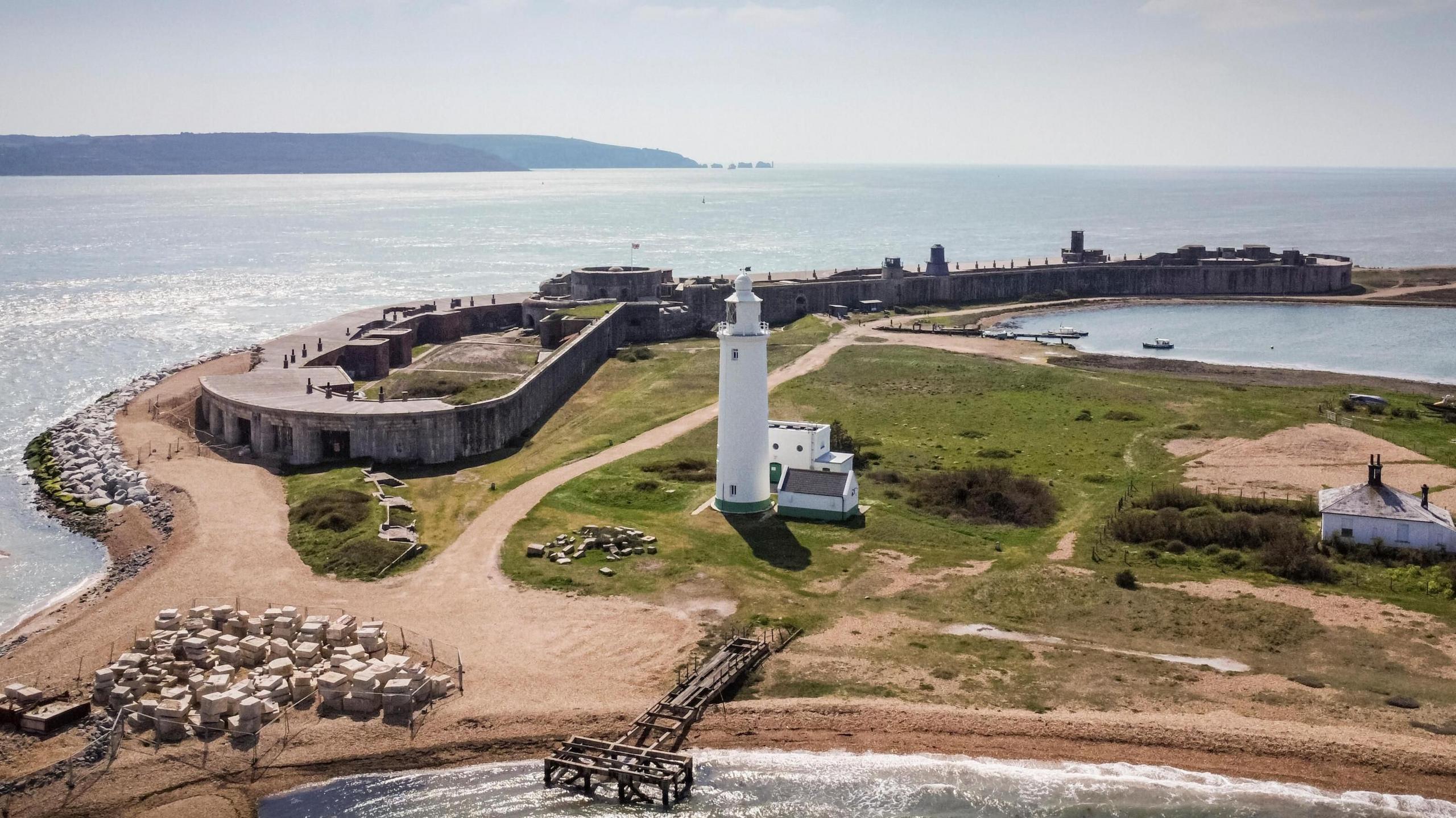 Hurst Castle, with its perimeter spanning the tip of the shingle bank, and a white lighthouse in the foreground, pictured at the tip of Hurst Spit.