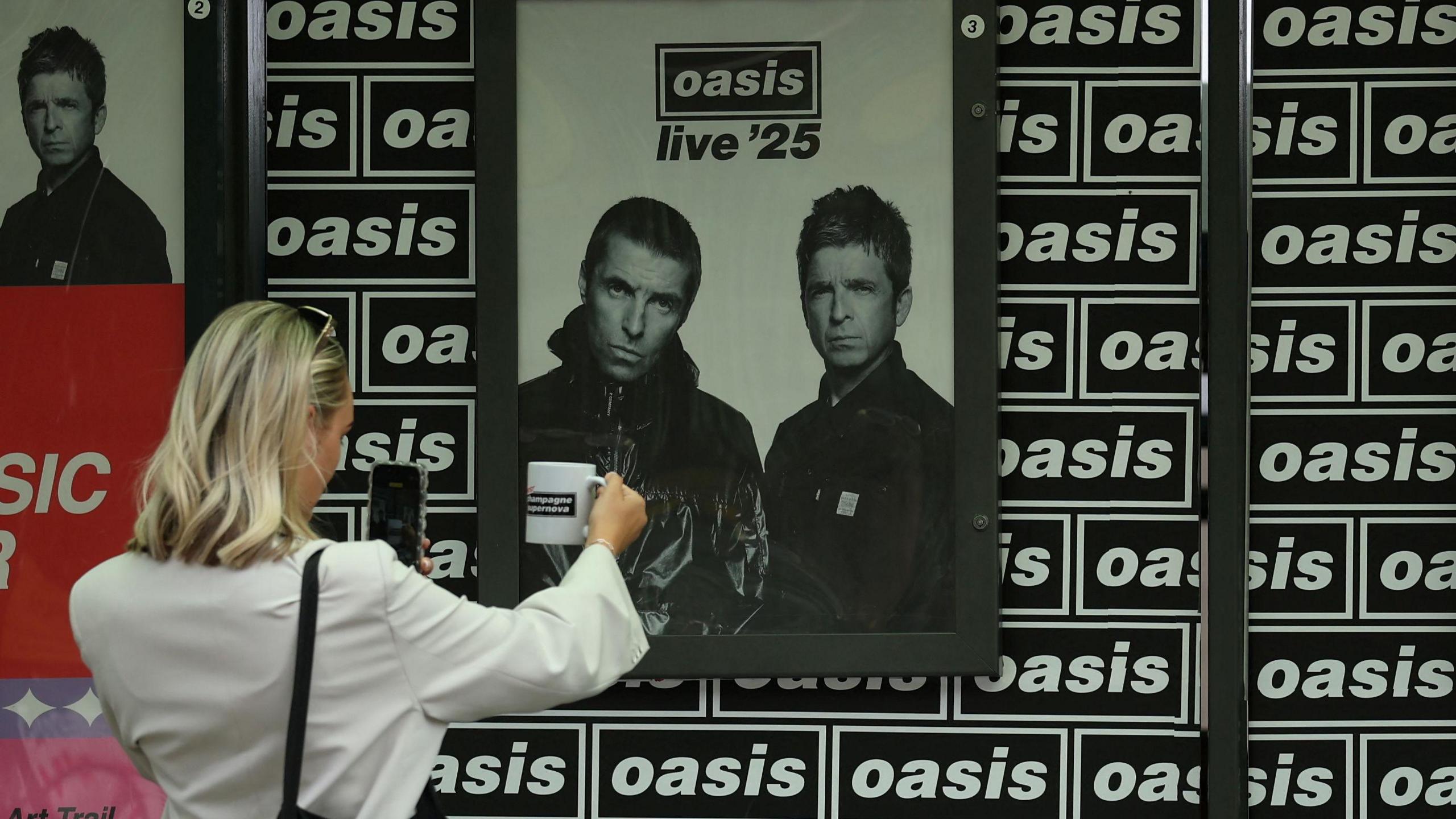An Oasis fan, wearing a white shirt and sunglasses on her head, holds an Oasis-branded cup up in front of her phone to take a picture with an image of Liam and Noel Gallagher and black and white Oasis-branded wallpaper