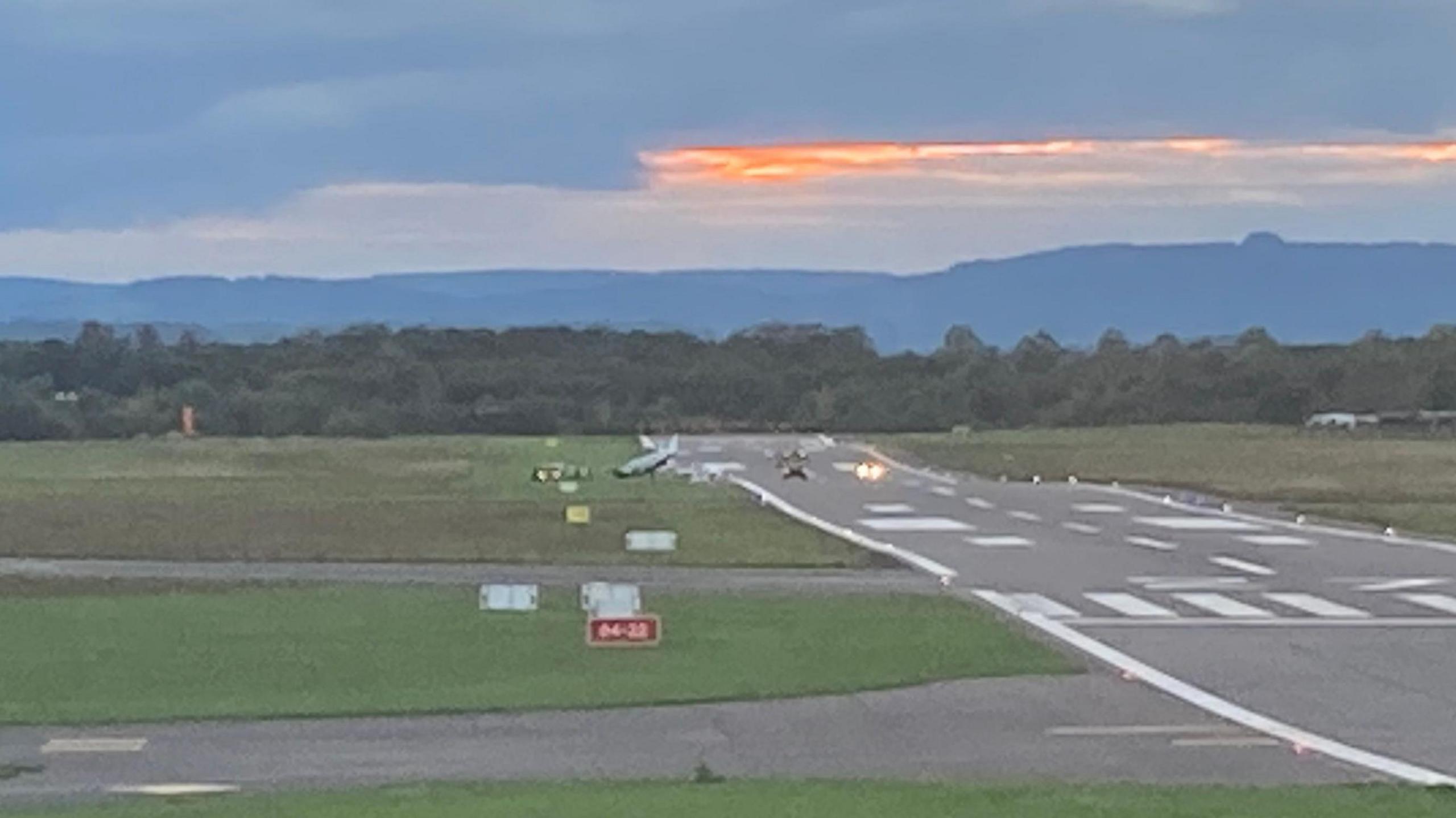 A picture of the runway at Gloucestershire Airport. The plane can been seen on the grass, with the nose slightly pointed down. At least two vehicles appear to be next to the plane. 