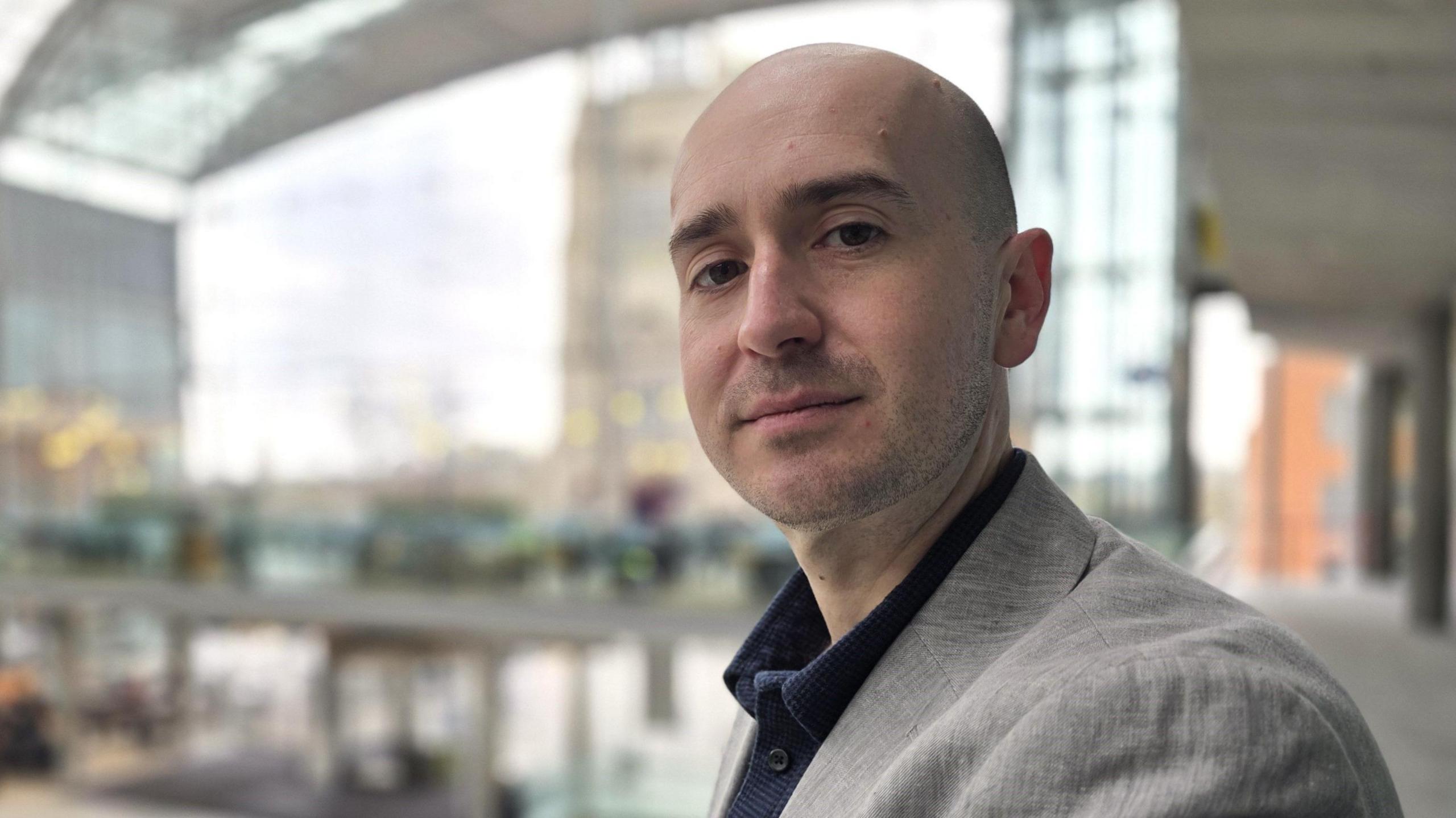 Ben Goldsborough who is standing inside The Forum in Norwich. He is looking directly at the camera and is wearing a grey suit jacket and dark shirt.