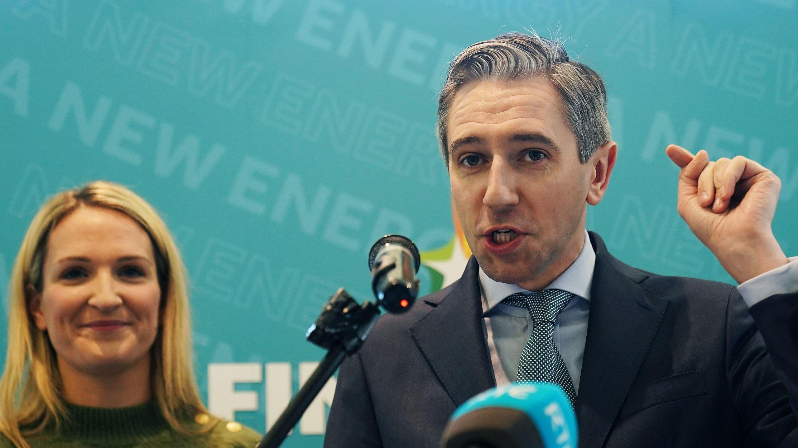 A woman with blonde hair standing beside a man with grey hair who is wearing a navy suit. There is a blue background behind them. 