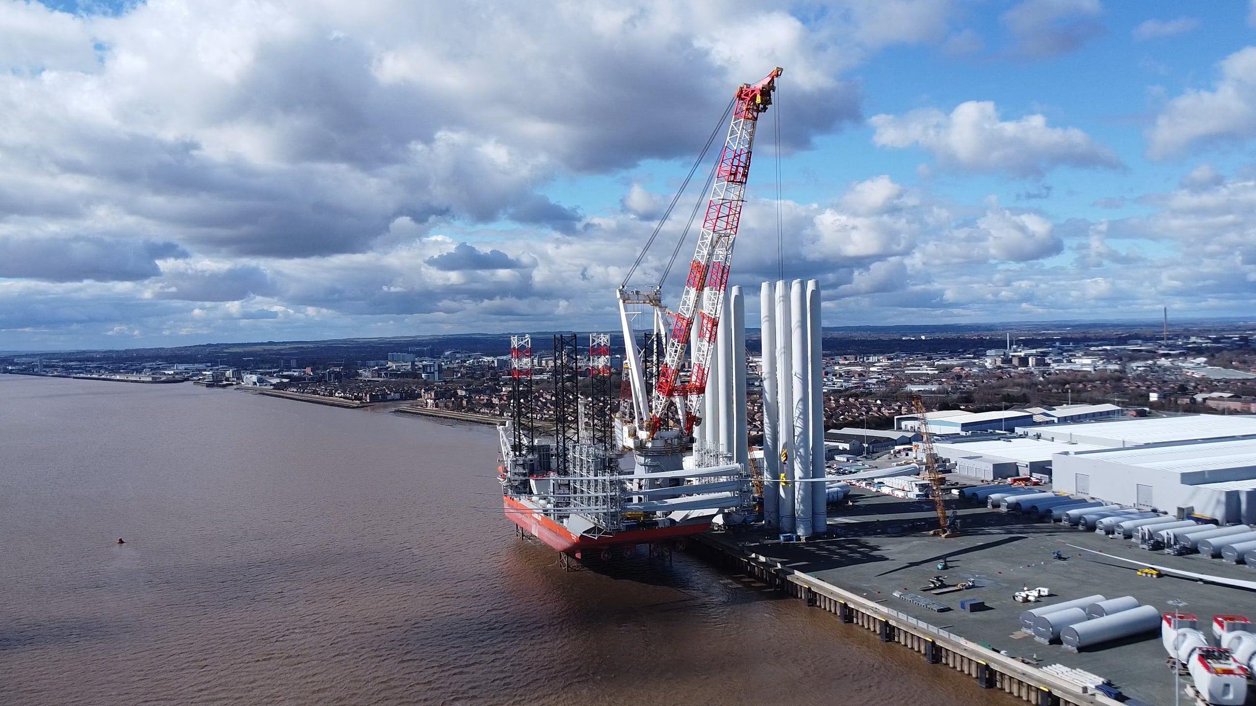 A large vessel is moored alongside a dock, next to a factory. It is loaded with components for wind turbines and cranes.