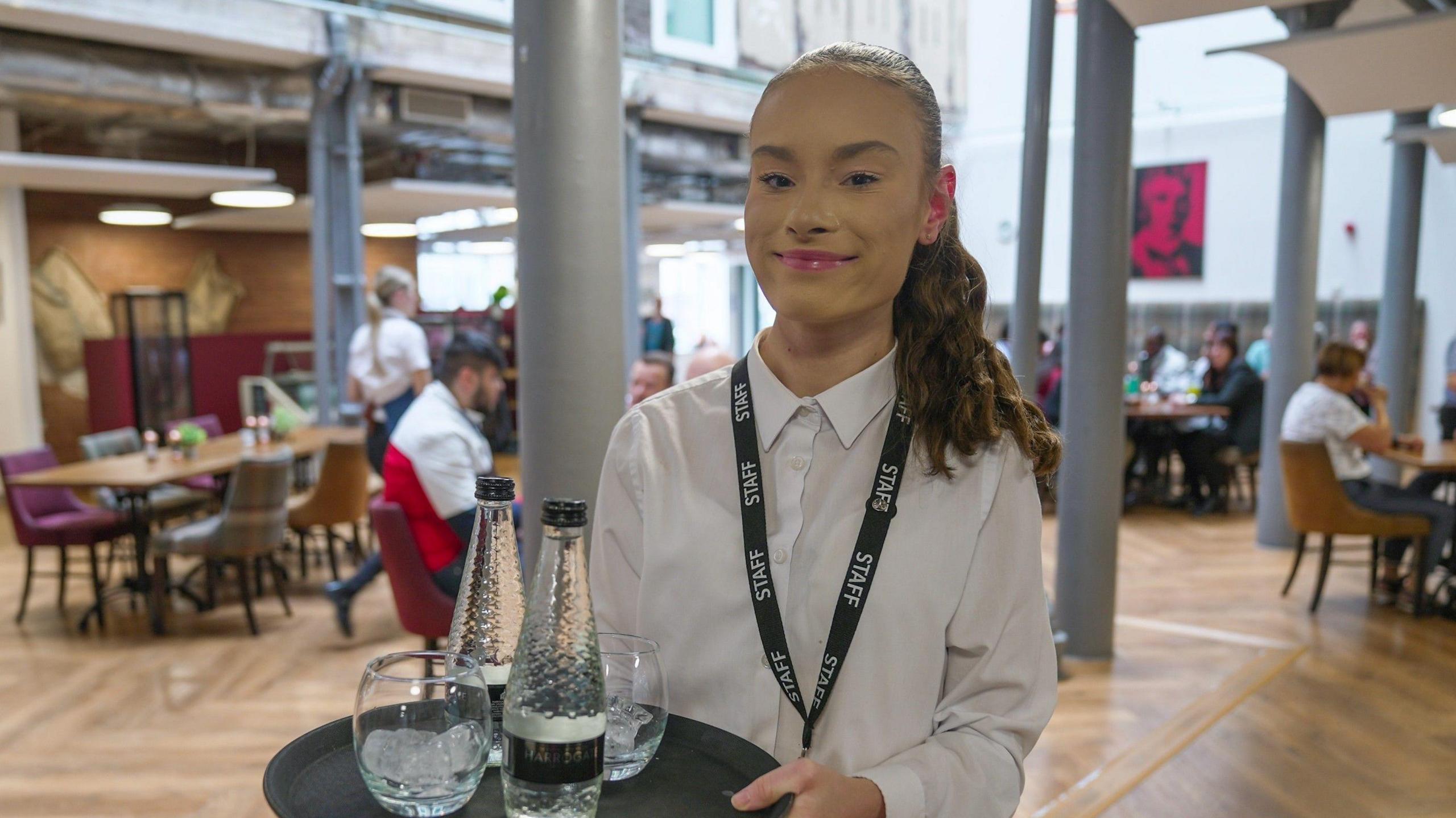 Isabel is smiling into the camera while holding a tray with two bottles of water and two glasses on it. She is at work at a cafe with people sat at tables in the background, and is wearing a smart white shirt buttoned up to her neck.
