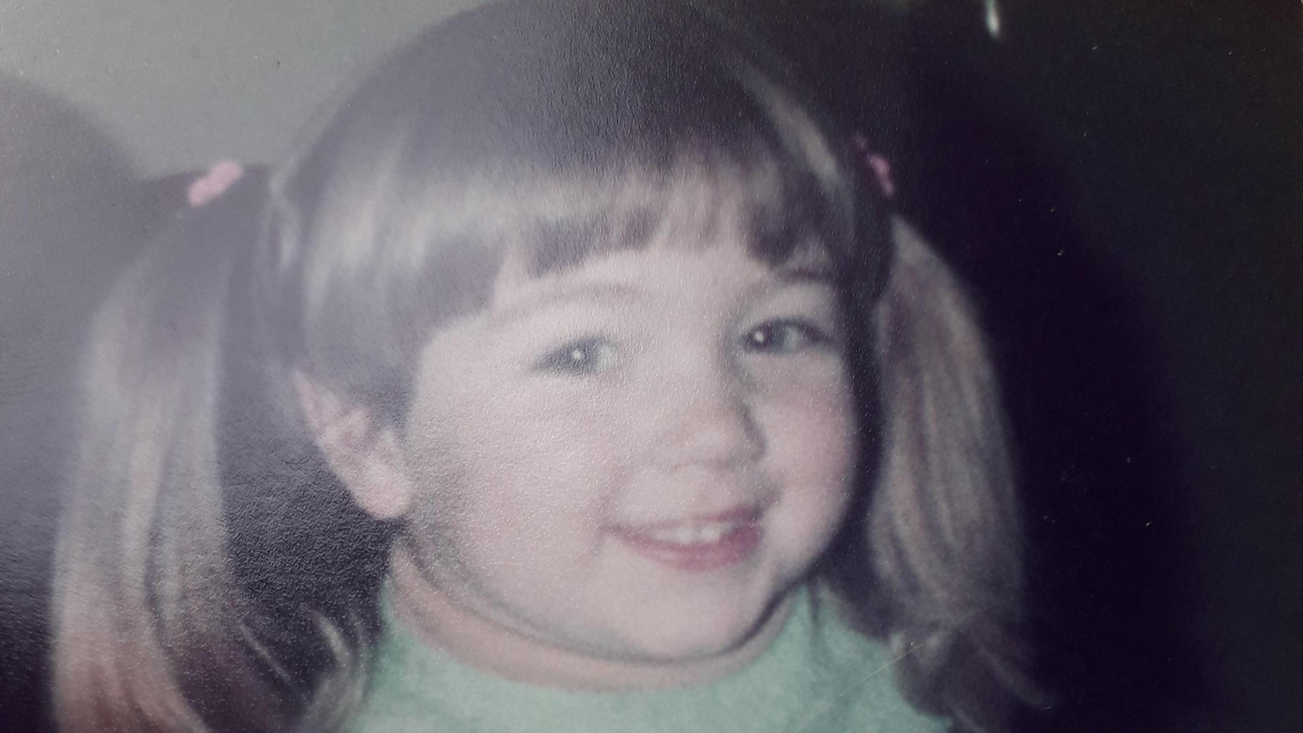 Steff when she was young with bunches in her brown hair. She is smiling at the camera and has a green jumper on.