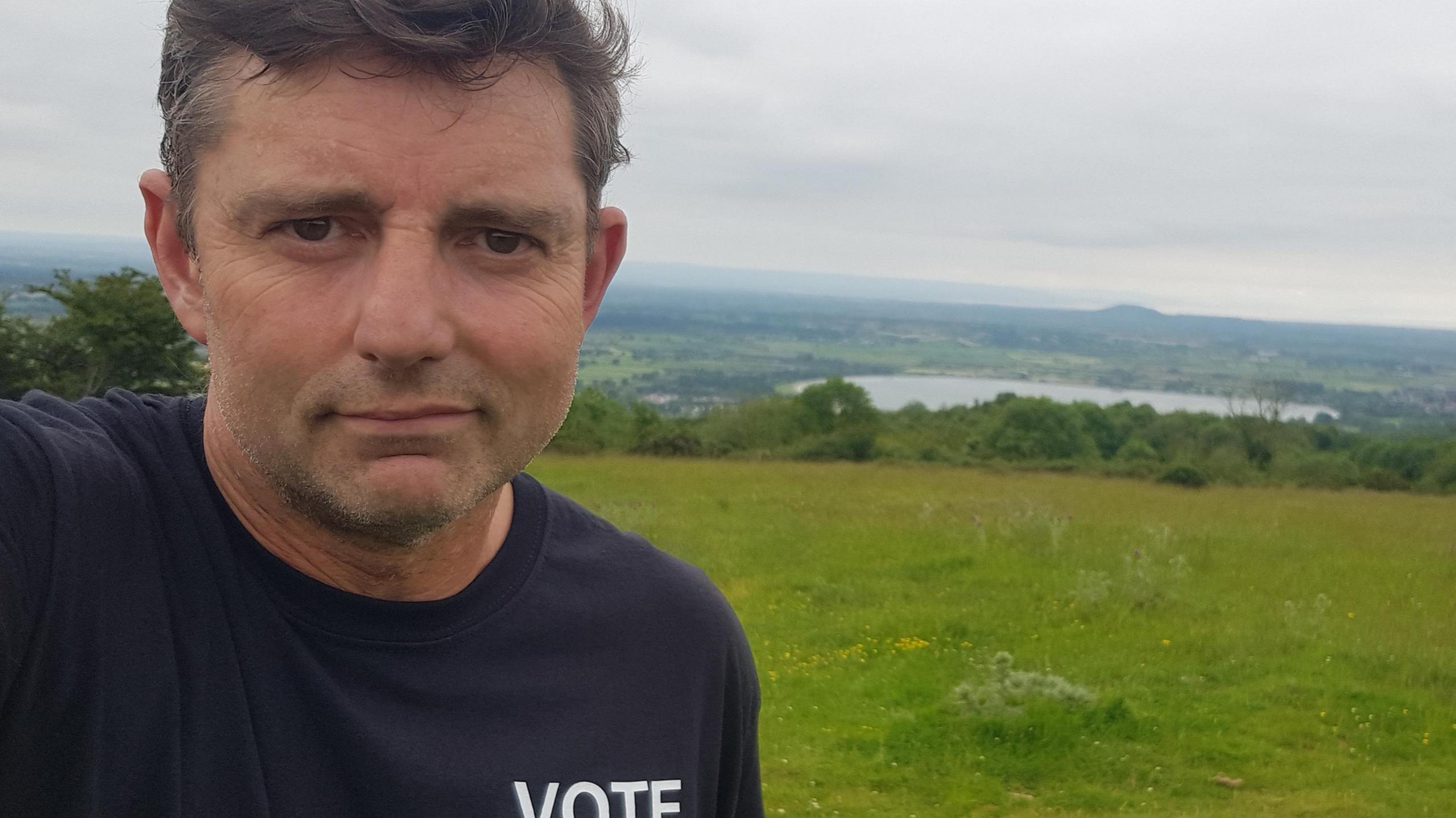 Craig Clarke wearing a blue top in a field overlooking a reservoir.