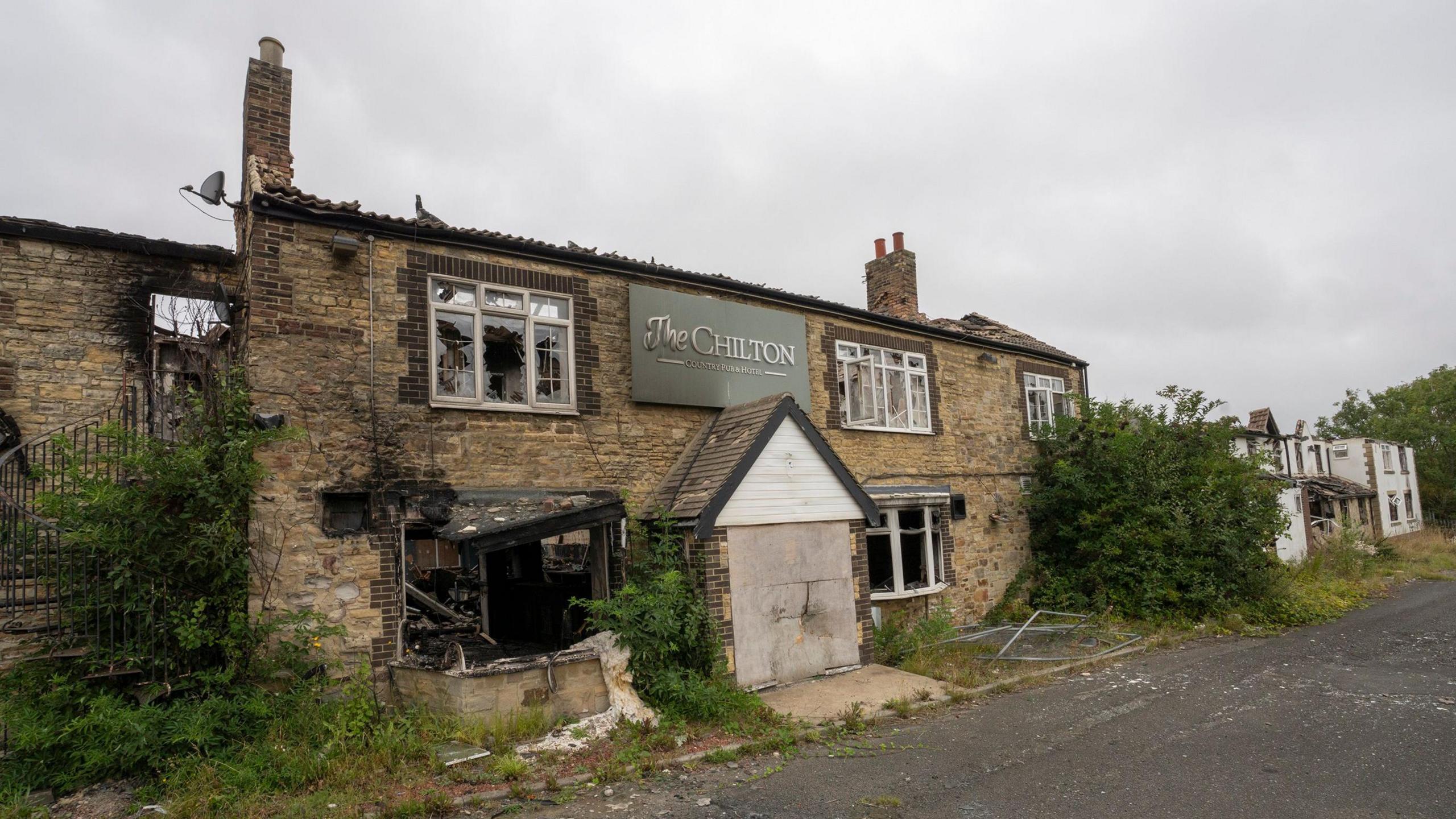 A stone building with two chimneys, and burnt out windows, and a boarded up door. A sign reads 'The Chilton' which has black marks on it from smoke. 